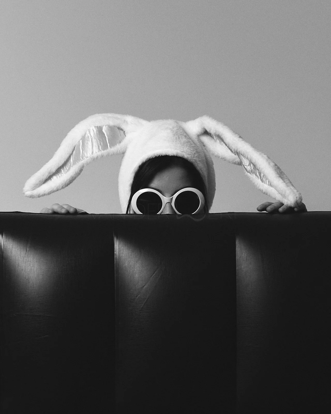 A person wearing large bunny ears and round sunglasses peeking over a black surface in a black-and-white photo.