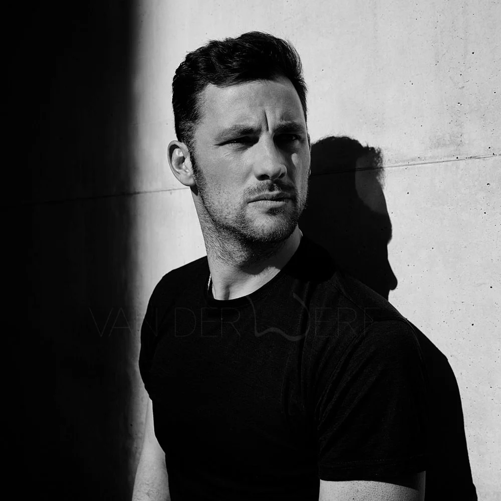 Black and white photo of a man with short dark hair and a beard, standing with back against a wall, casting a shadow on the wall, wearing a black shirt.