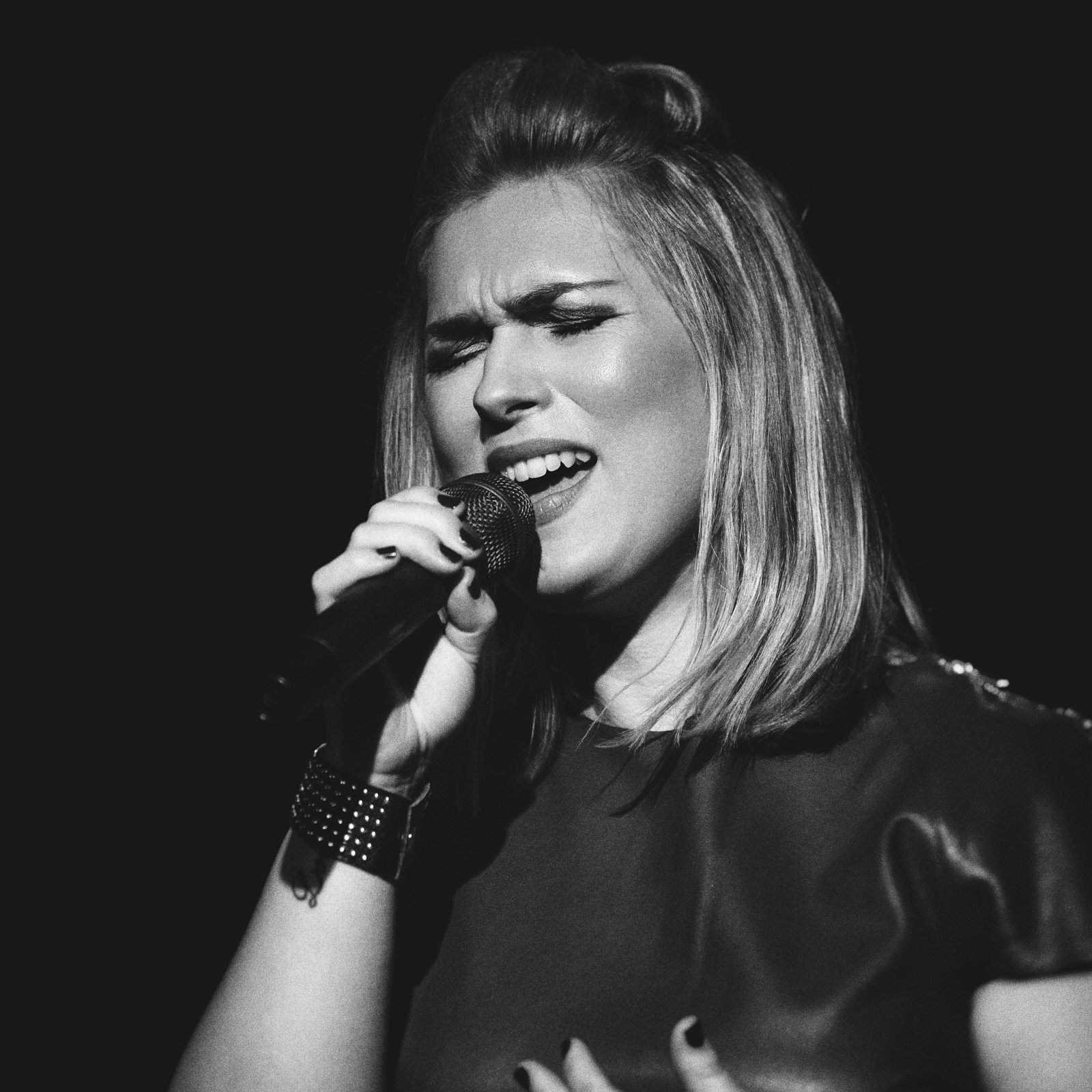 A woman singing passionately into a microphone, eyes closed, with a black background.