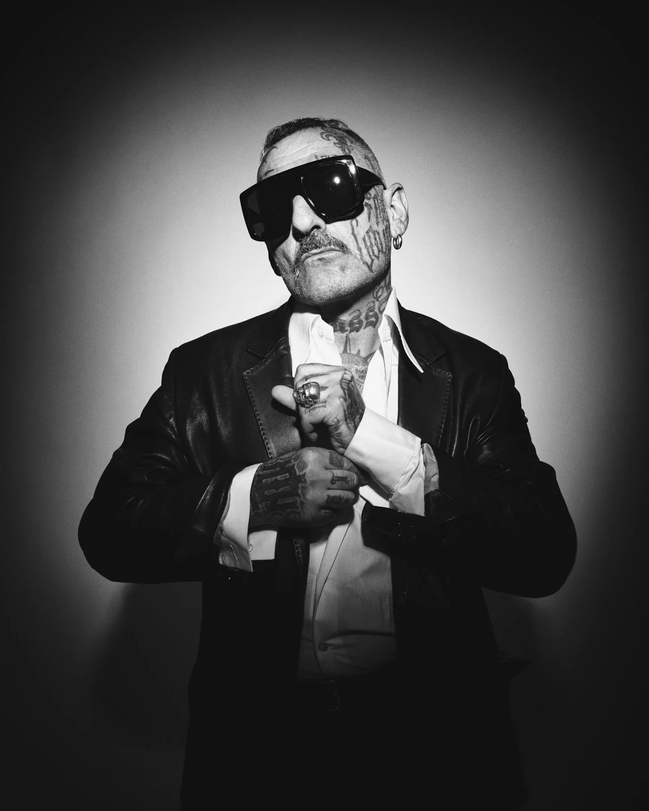 Black and white portrait of a tattooed man wearing large sunglasses, a leather jacket, and a white shirt, standing against a plain background.