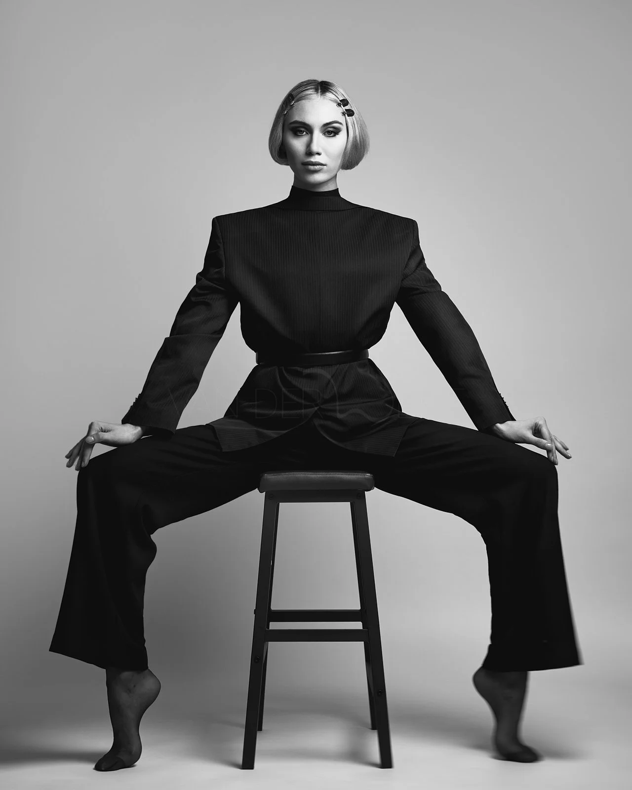 A black and white photo of a woman sitting on a stool with her legs spread apart. She is wearing a black outfit with long sleeves and has her hair styled with clips.