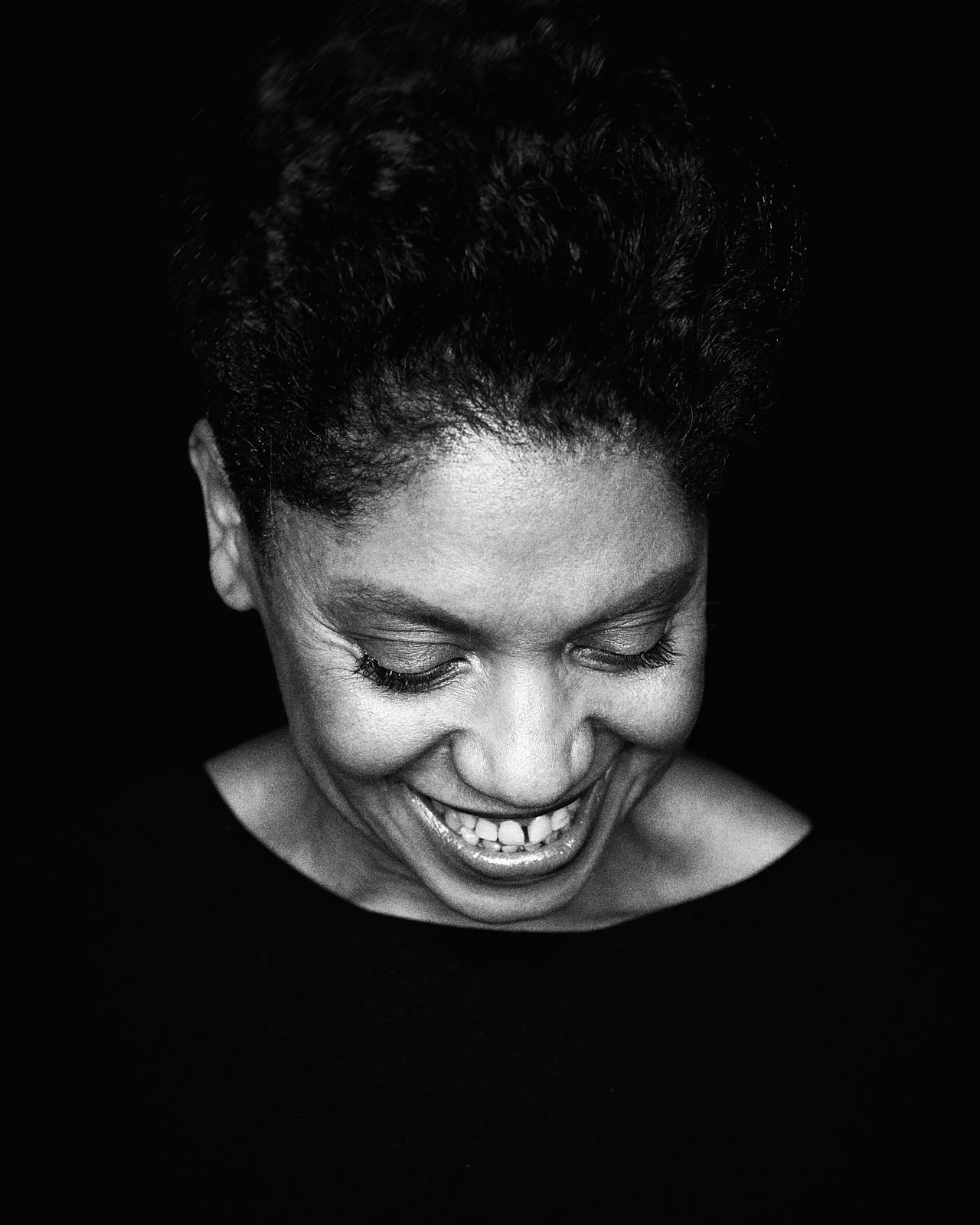 Black and white photograph of a woman smiling with her eyes closed, head tilted downward, dark background.