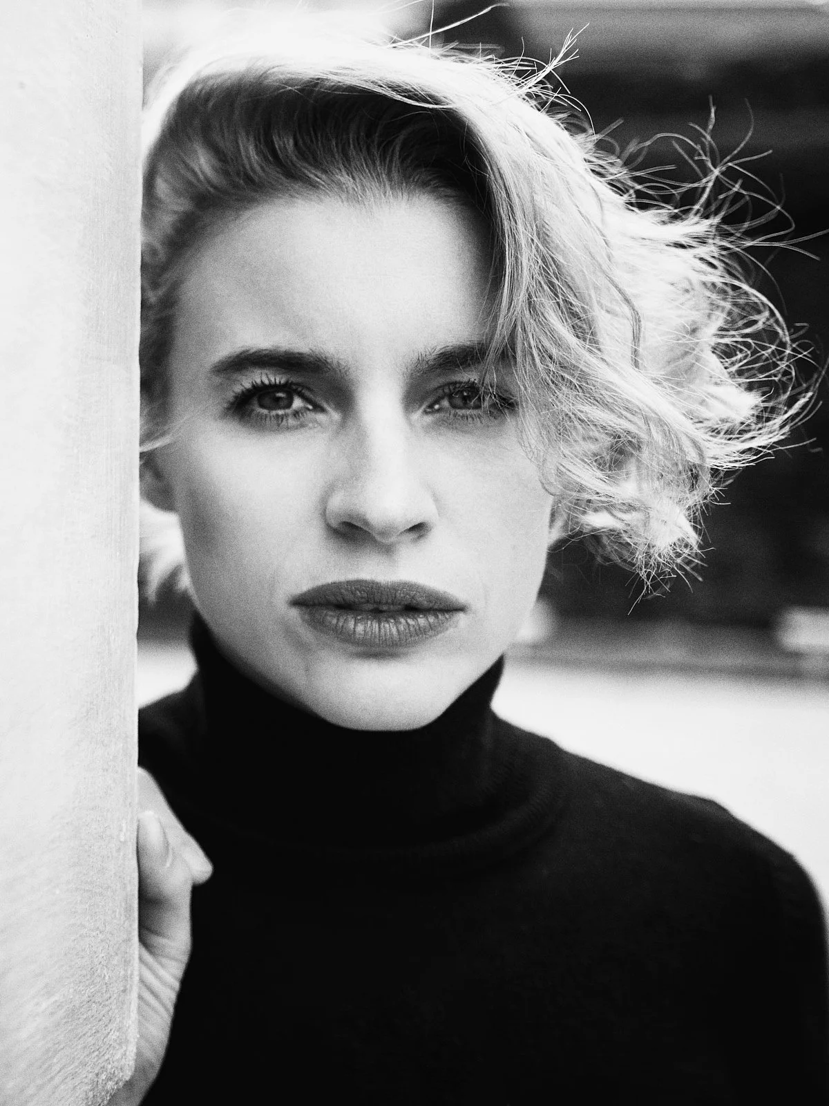 A black and white close-up portrait of a woman with short, wavy hair and intense eyes, wearing a black turtleneck, partially obscured by a white surface she is resting her hand on.