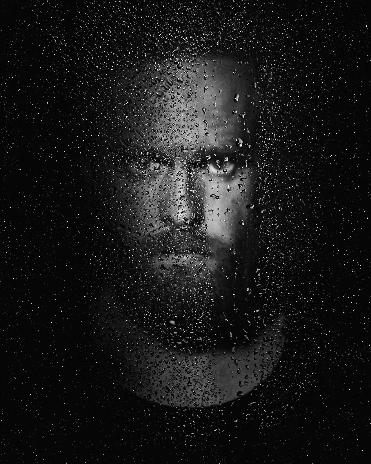 Black and white photo of a man with glasses looking through a rain-covered glass surface.