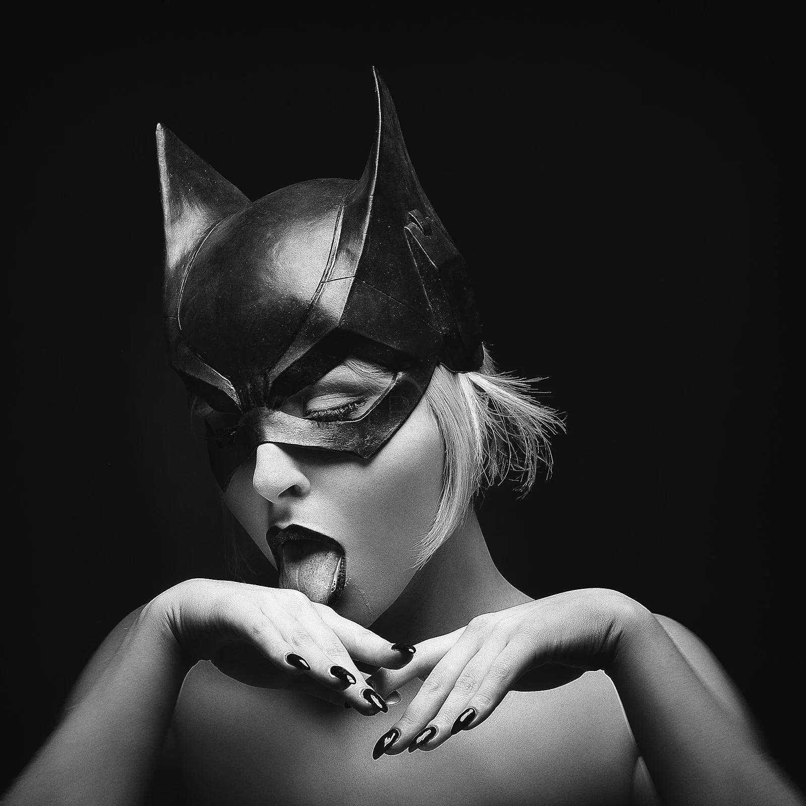 Black and white photo of a woman wearing a catwoman-style mask, sticking out her tongue, with her hands near her chin and short blonde hair.