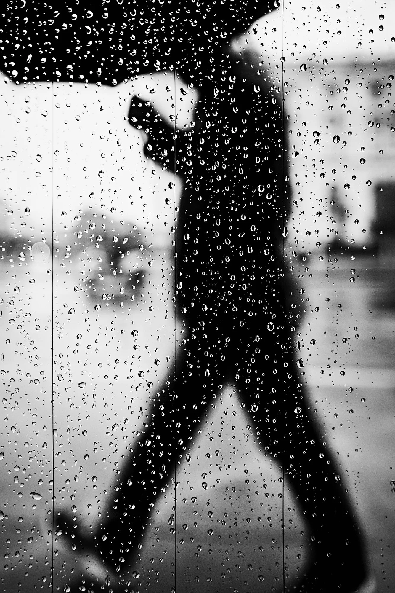 Silhouette of a person walking outside, viewed through a rain-covered window with water droplets on the glass, black and white photograph.