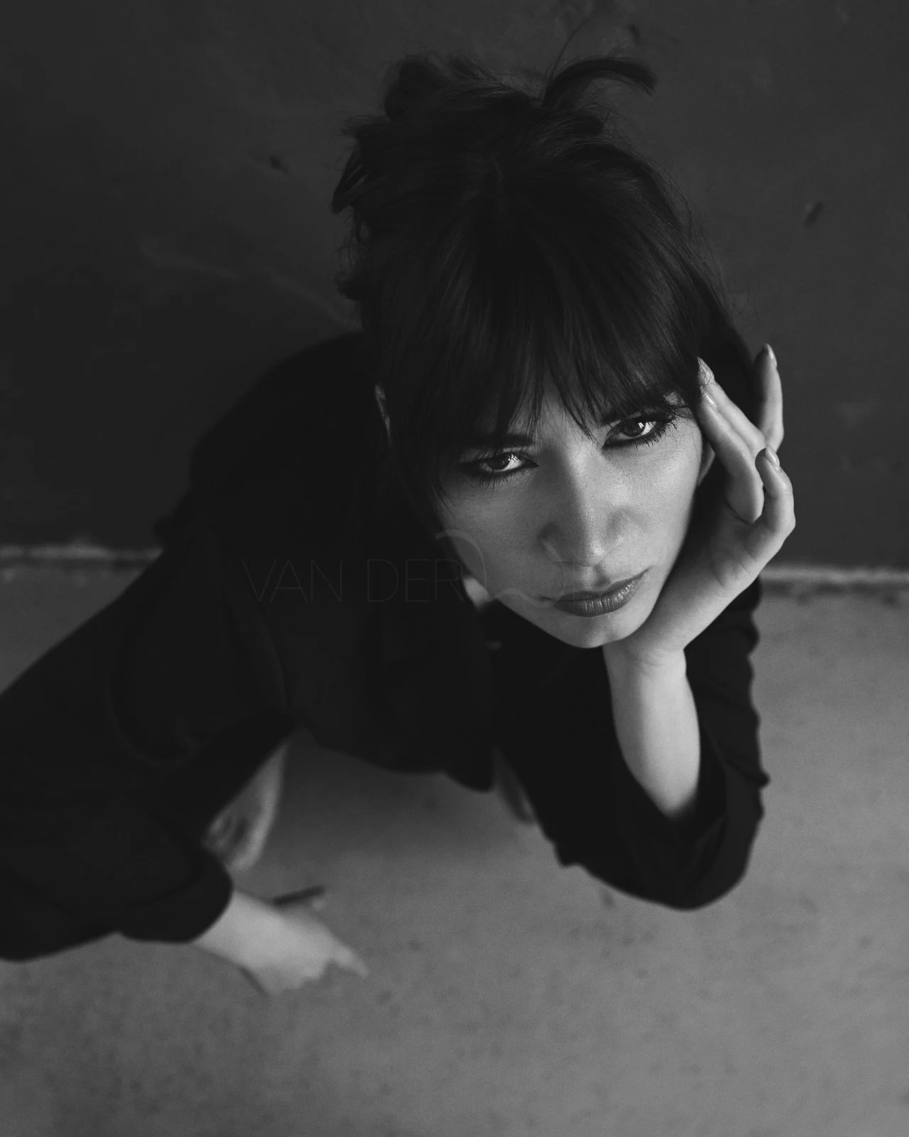 Black and white photo of a woman with short dark hair, looking up at the camera with an intense gaze, her hand resting on her cheek, standing on a sandy surface.