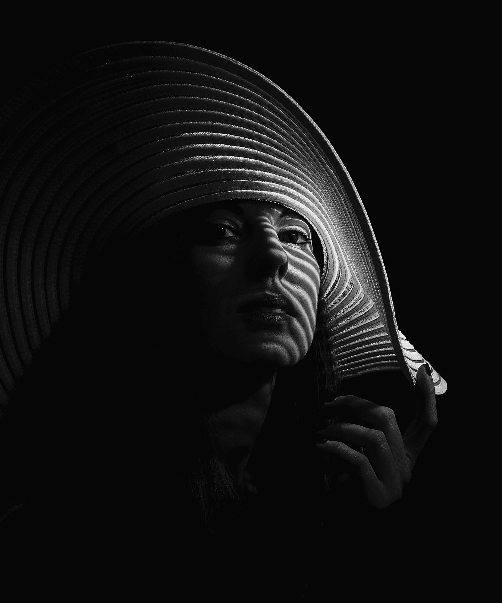 A woman wearing a large, wide-brimmed hat with a striped pattern, partially illuminated, with sunlight casting shadow lines across her face, against a dark background.