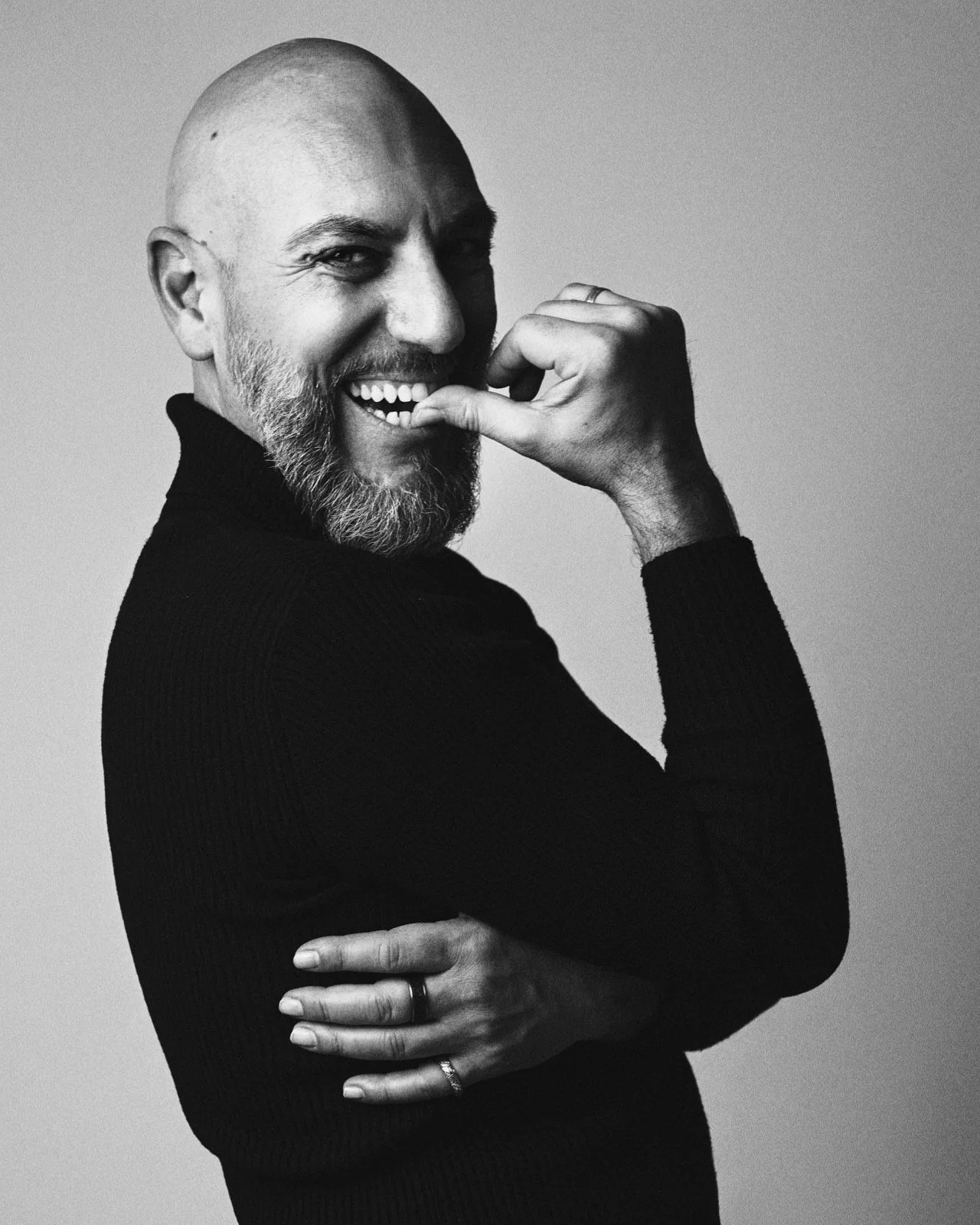 A black and white portrait of a smiling, bald man with a beard, wearing a black sweater, with one arm crossed and the other hand near his mouth.