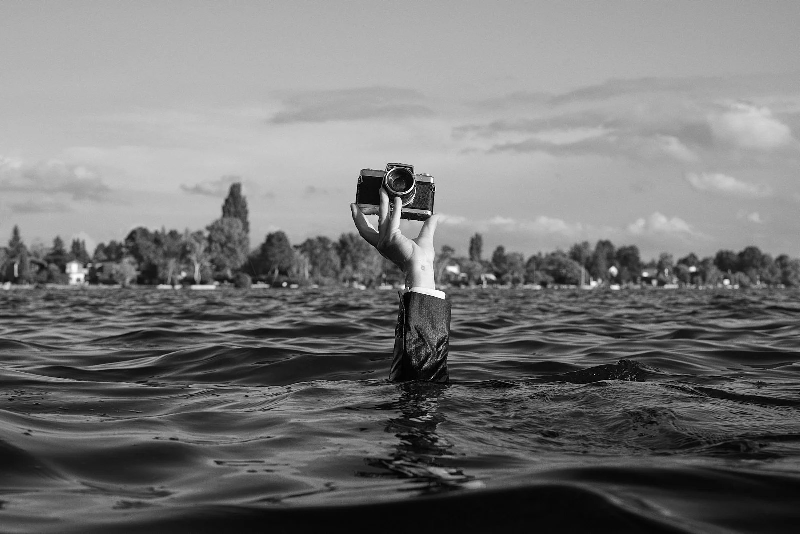 A person in a suit submerged in water up to their arm, holding a camera above their head with the shoreline visible in the background.