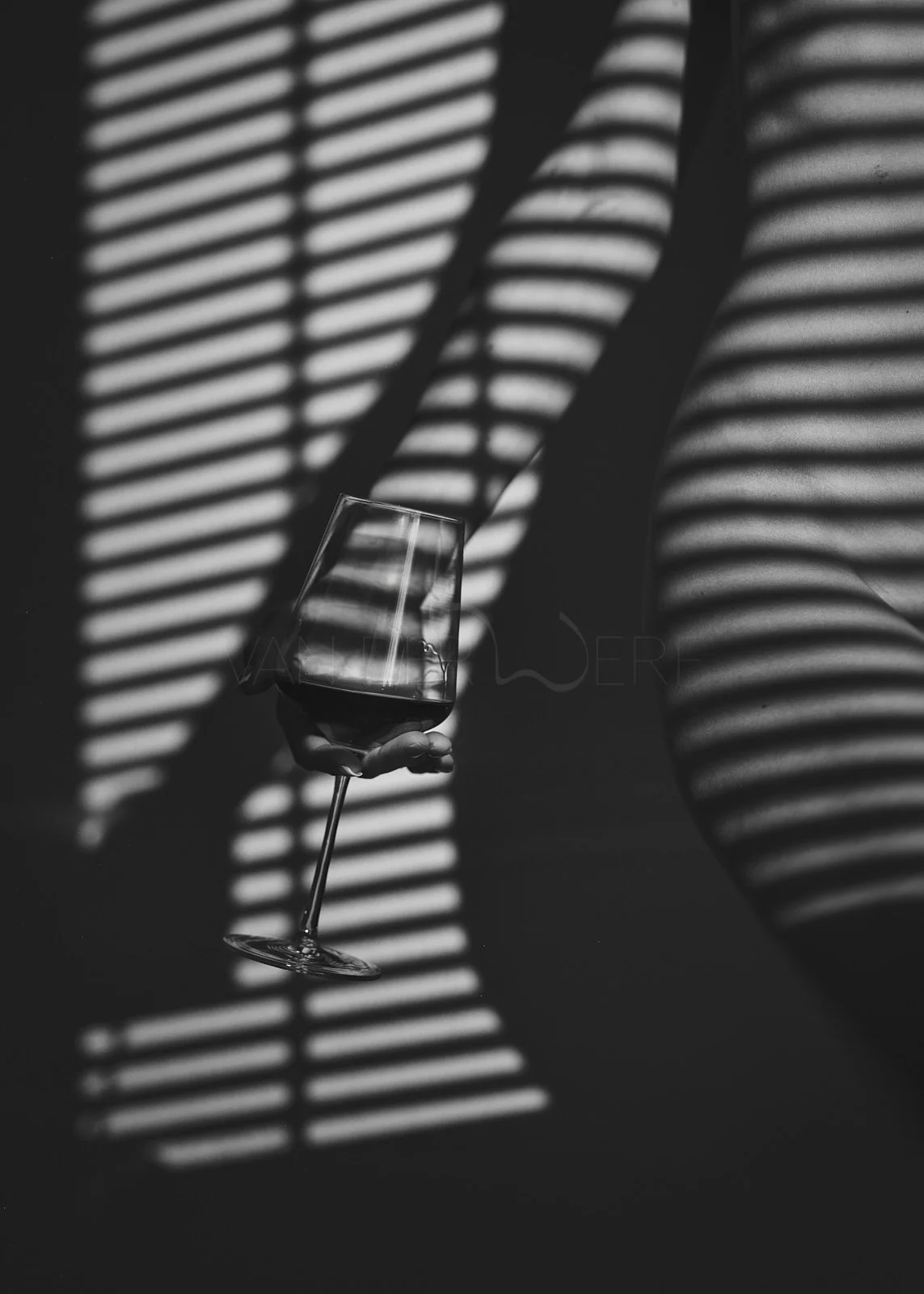 A person holding a wine glass with dark liquid, with striped shadows cast by window blinds creating a pattern on the person's body and the background, in black and white.
