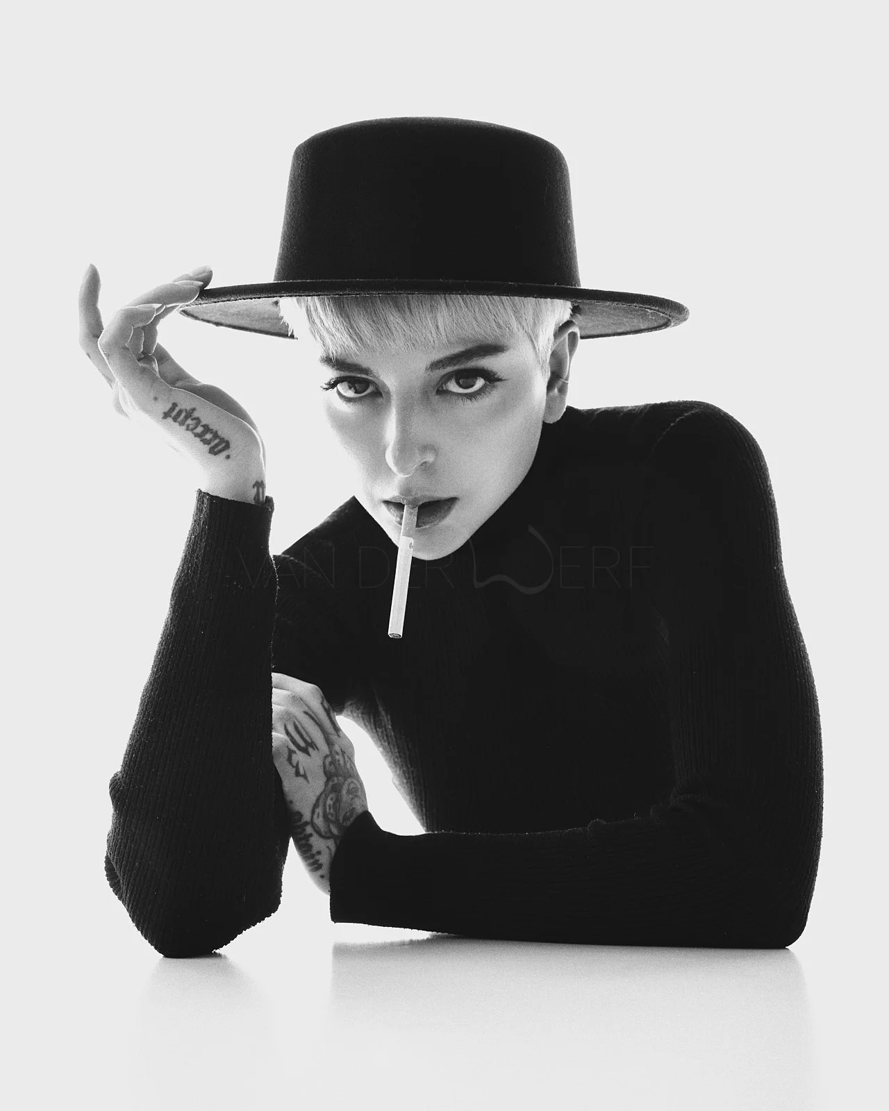 Black and white photo of a woman with short blonde hair wearing a wide-brimmed hat and a dark sweater, looking directly at the camera with a lollipop in her mouth, holding her hat with one hand.