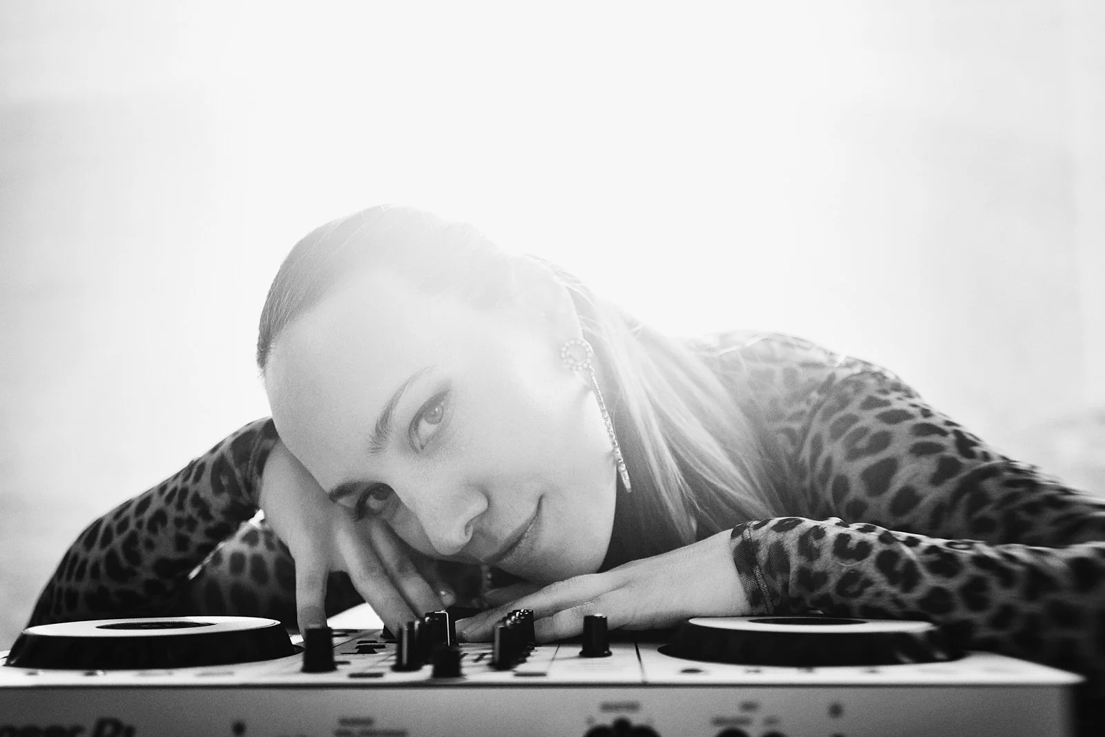 A woman with leopard print shirt resting her head on a DJ mixer, looking at the camera with a smile, illuminated by bright backlight.