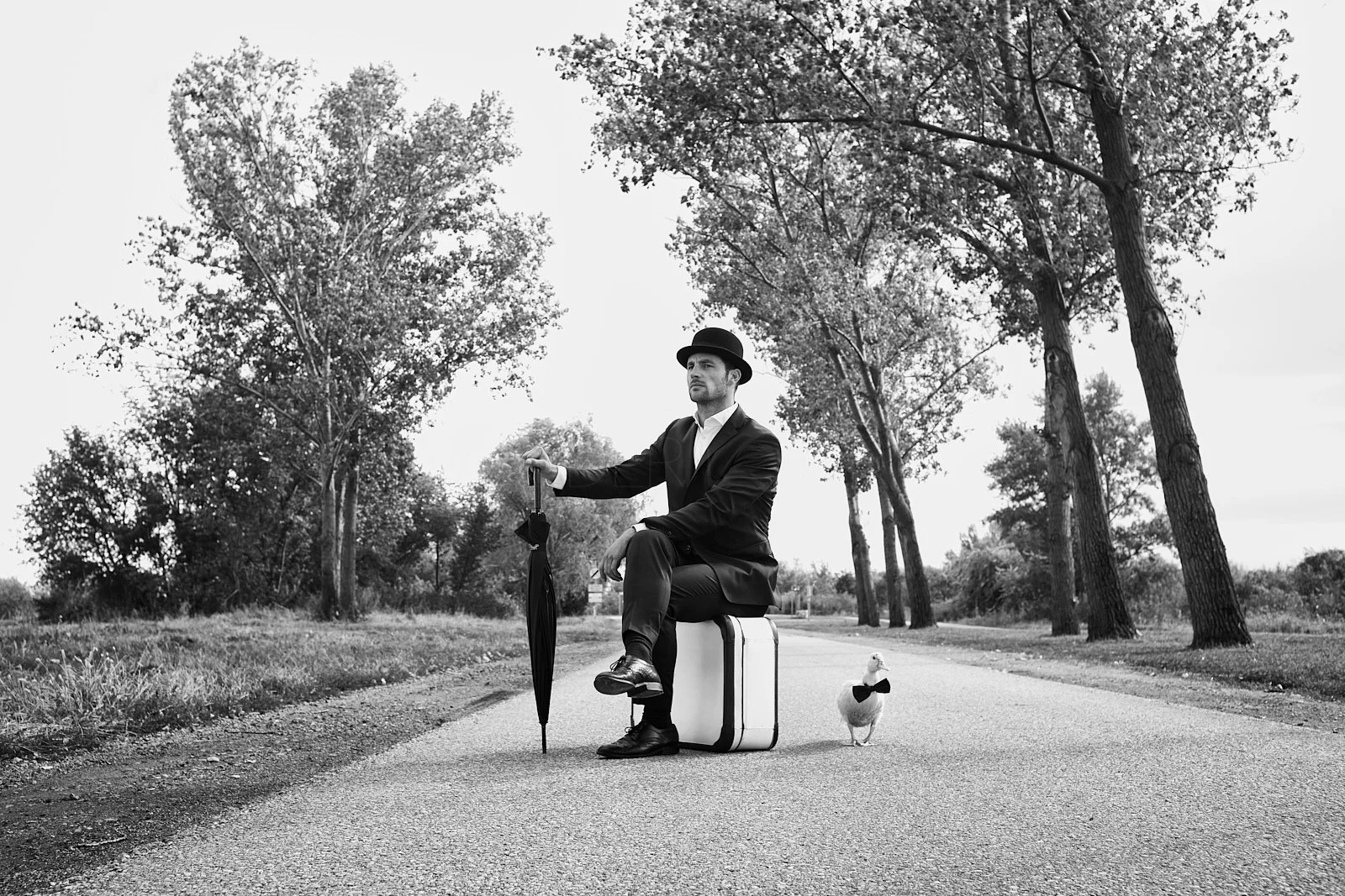 A man dressed in a suit and hat sitting on a suitcase next to a goose in a rural area with tall trees, holding an umbrella, in black and white.
