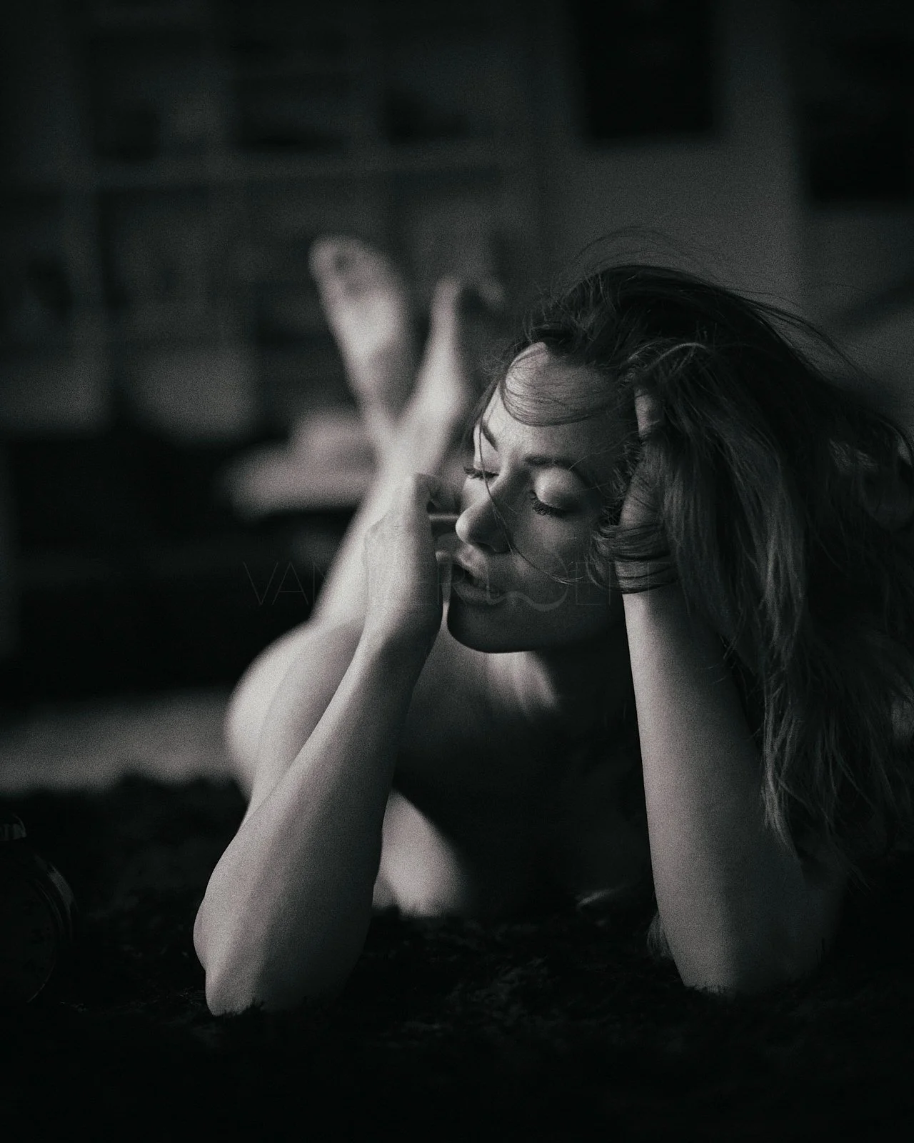 A woman is lying on her stomach on a dark surface with her eyes closed, resting her head on one hand and the other hand on her forehead, with her legs crossed in the background.