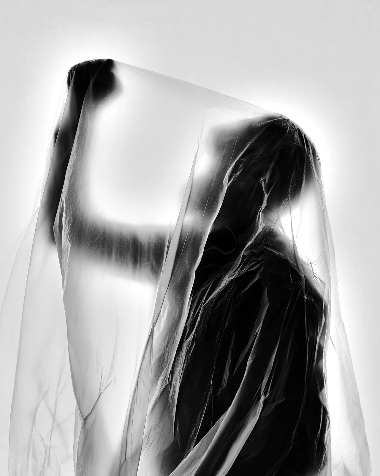 Black and white abstract photograph of a person wrapped in translucent plastic or fabric, with the outline of a face visible through the material.