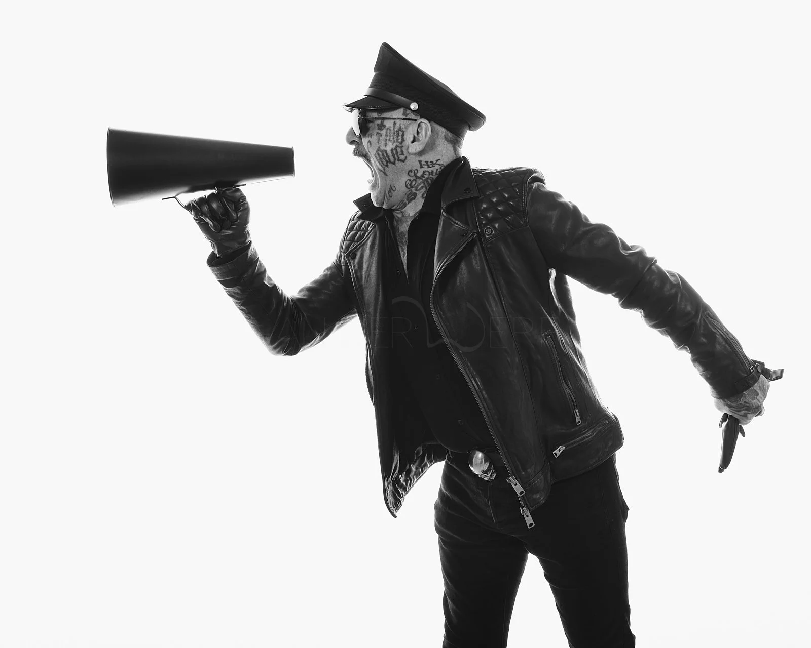 Black and white image of a tattooed man in a leather jacket wearing a police cap, shouting into a megaphone, with a gun in his other hand.