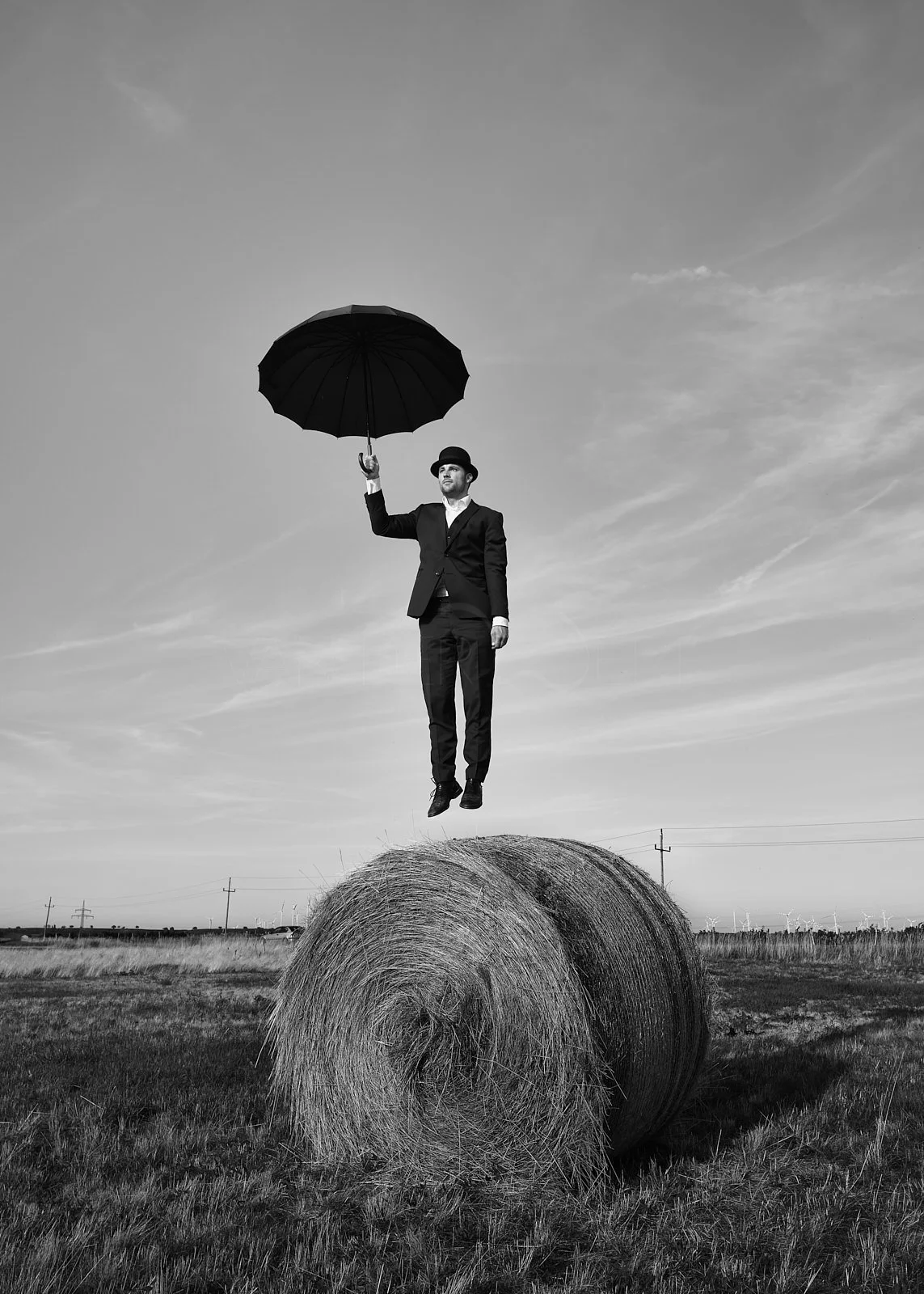 A man dressed in a suit and bowler hat, holding an umbrella, appears to be floating above a hay bale in an open field with wind turbines in the background, captured in black and white.