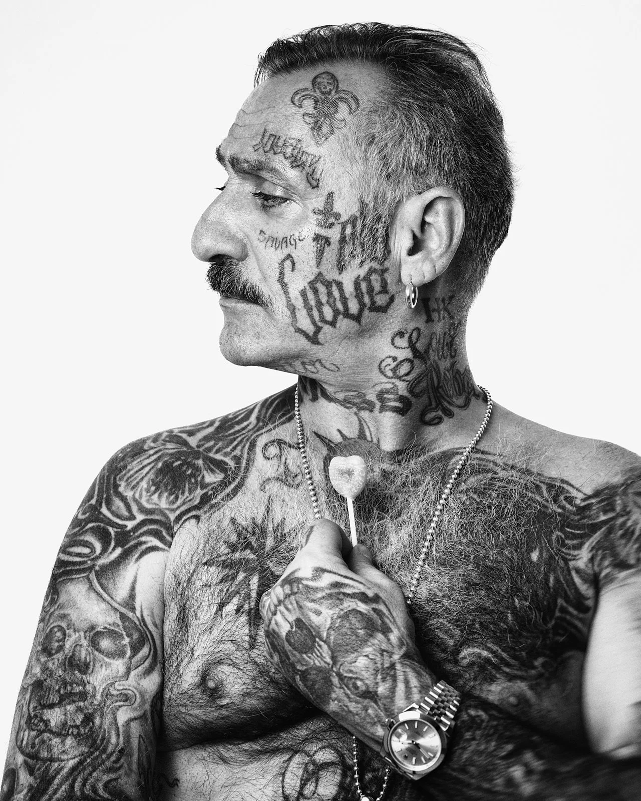 Black and white portrait of a shirtless man with numerous tattoos on his face, neck, and arms, holding a lollipop with a skull design. He has a mustache, earrings, a watch, and a necklace.