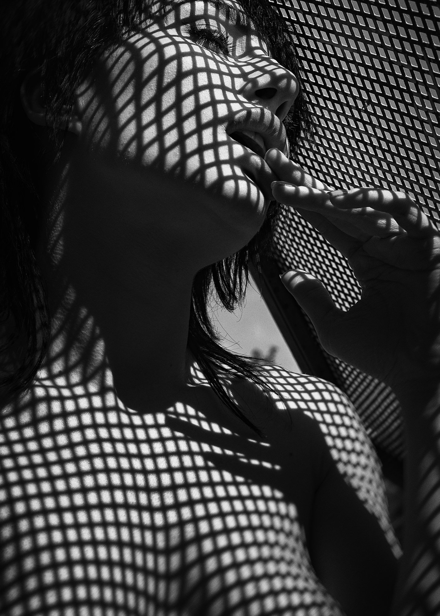 Black and white photo of a woman with sunlight casting a grid shadow on her face and body, with her hand near her face.