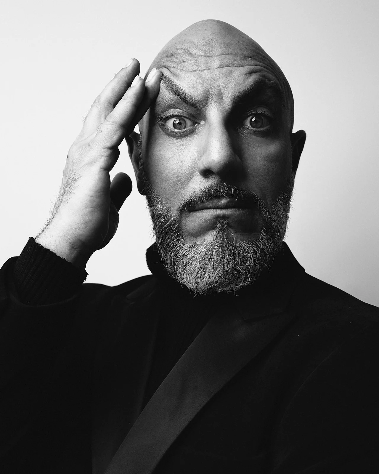 Black and white portrait of a bald man with a beard, looking introspective with one hand touching his forehead, wearing a dark blazer.
