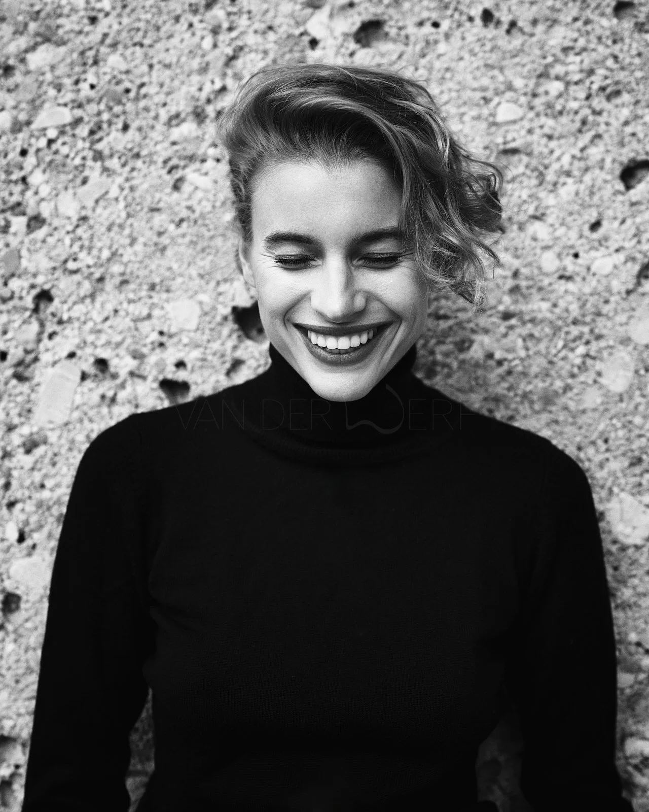 Black and white photo of a woman with short, wavy hair wearing a black turtleneck, smiling with eyes closed, standing against a rocky background.