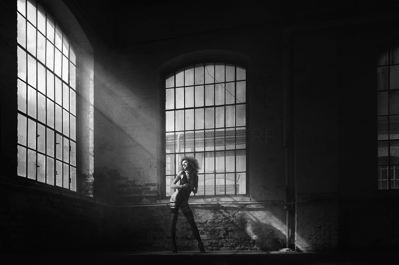Black and white photo of a woman with curly hair in lingerie and stockings, standing in front of large industrial windows with sunlight streaming in, in an abandoned industrial space.