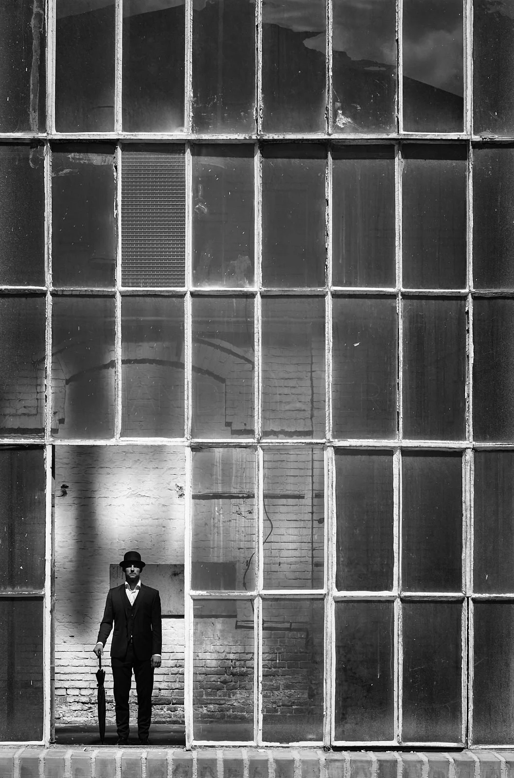 A man dressed in a suit and hat holding an umbrella, standing in front of a large industrial-style window with divided glass panes, a brick wall background, in black and white.