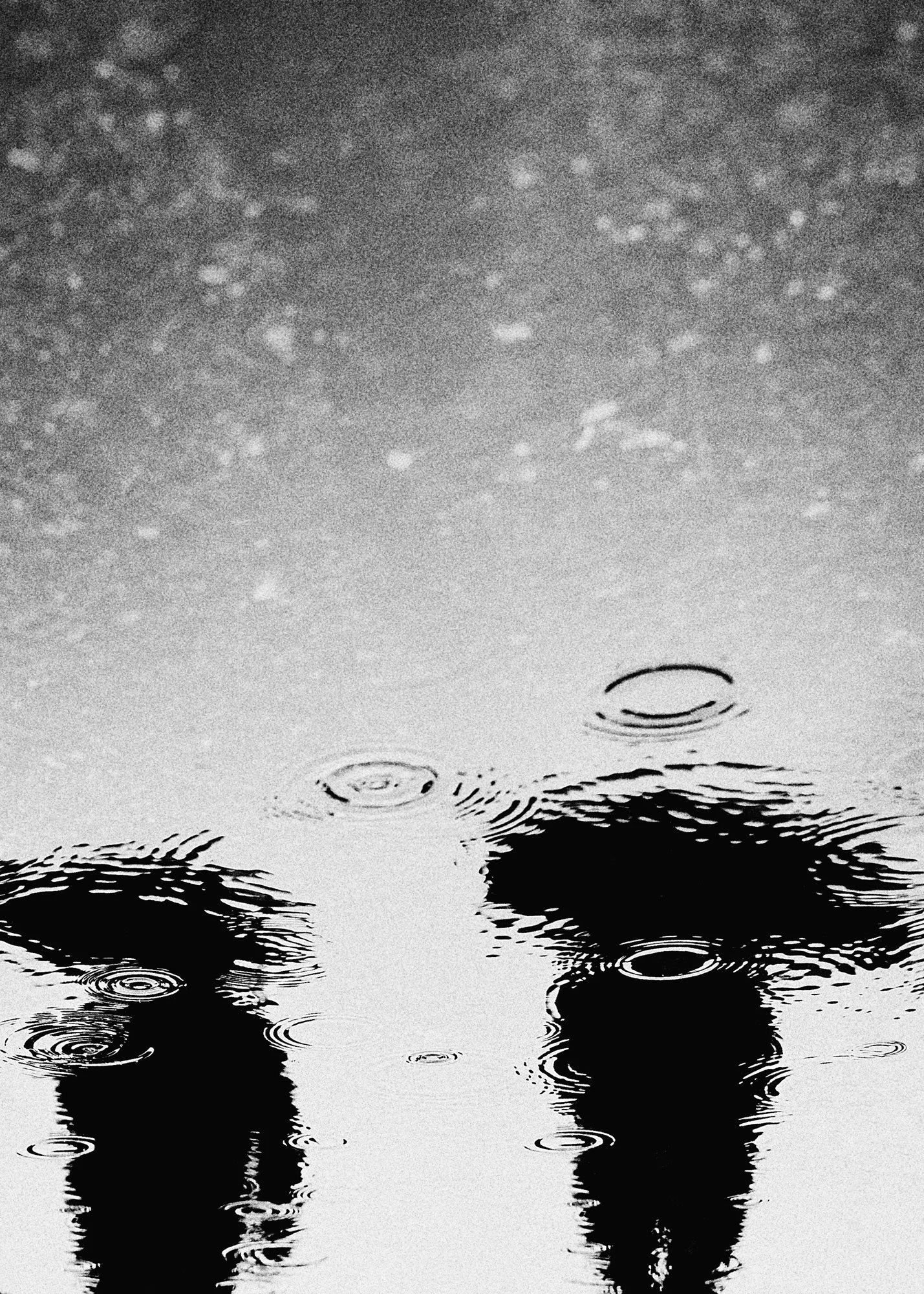 Black and white photograph of two people walking in the rain, with ripples on the water's surface and raindrops falling.
