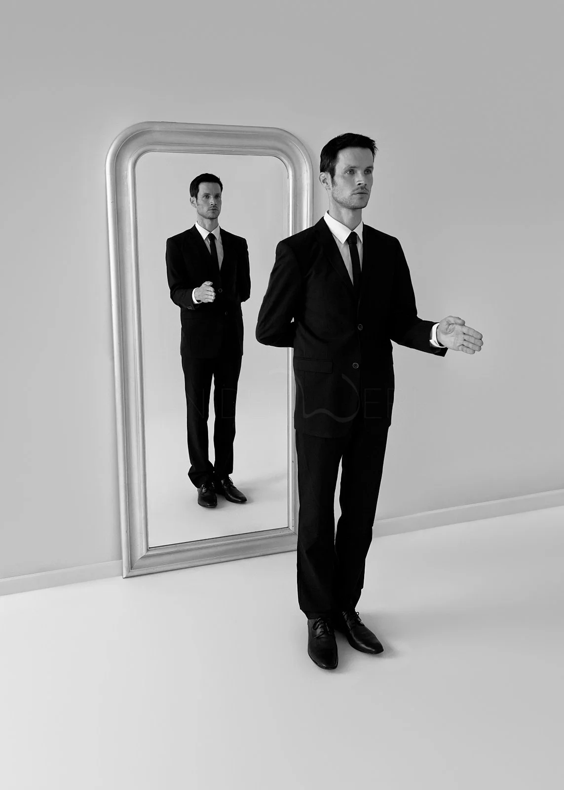 A man in a black suit and tie standing in front of a mirror, with his reflection visible behind him.