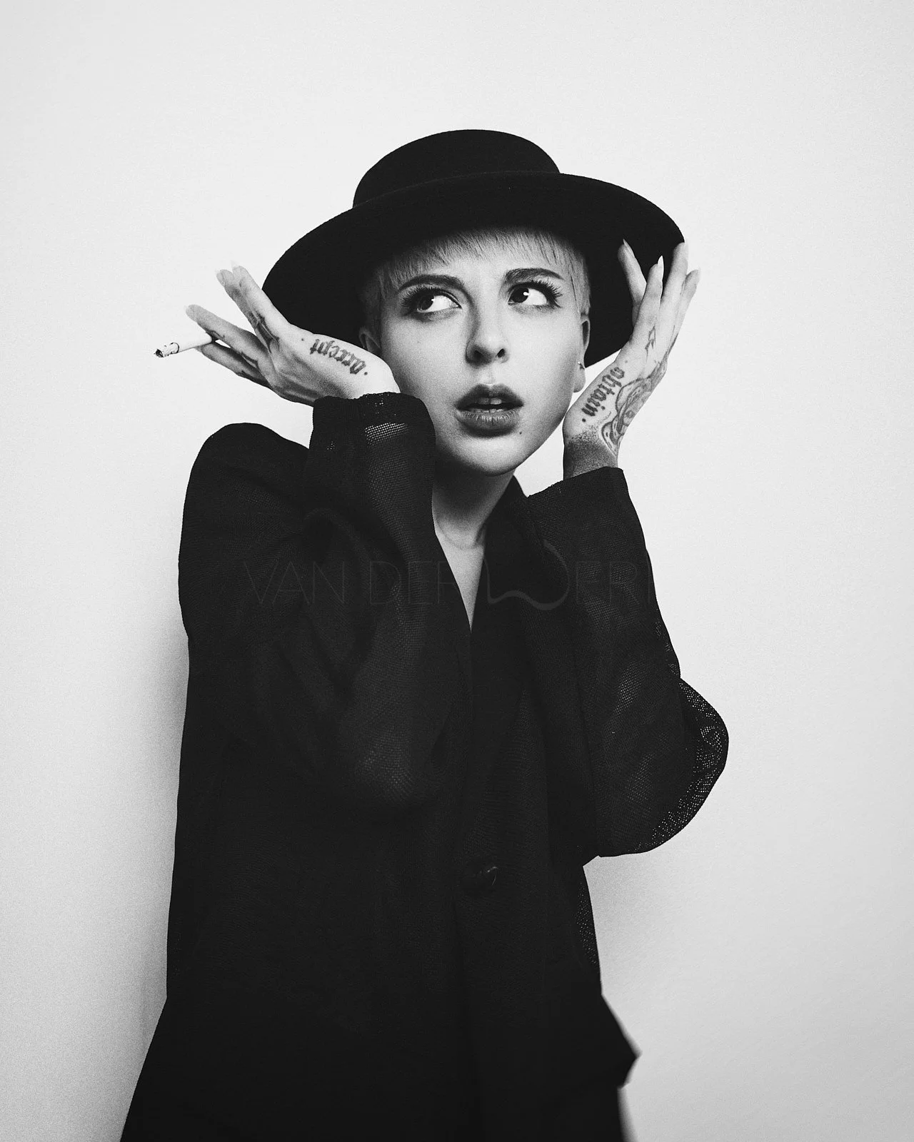 Black and white photo of a woman with short hair, wearing a wide-brimmed hat and dark clothing, holding her hands up to her face with tattoos visible on her fingers and wrists.