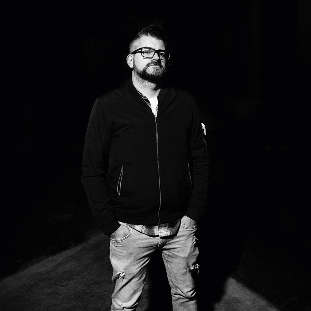 A man with glasses, a beard, and a mohawk hairstyle standing outdoors at night, wearing a black jacket, plaid shirt, and ripped jeans.