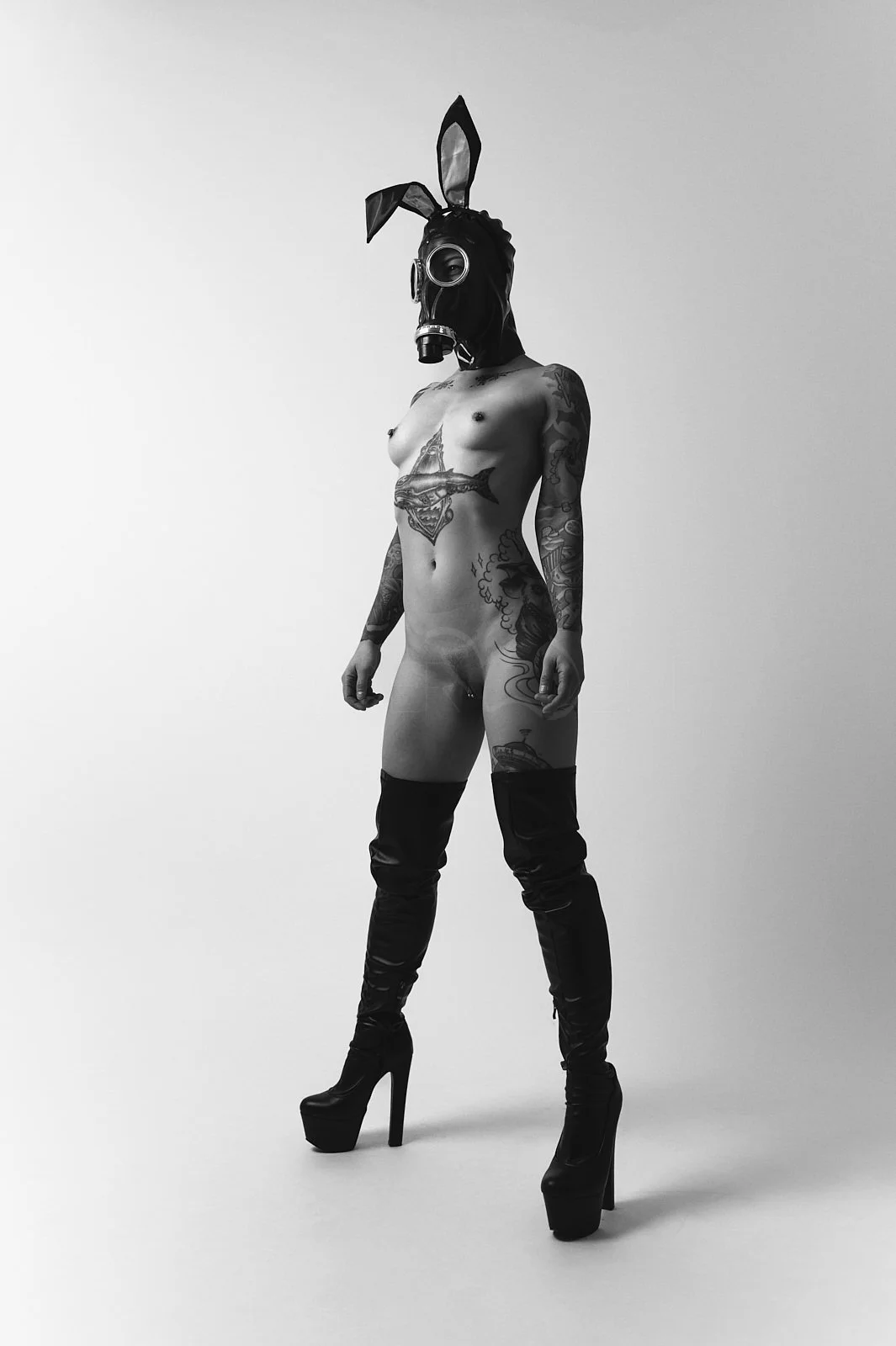 Person standing nude wearing a black bunny mask, high-heeled boots, and tattoos on their body in a minimalist studio setting.