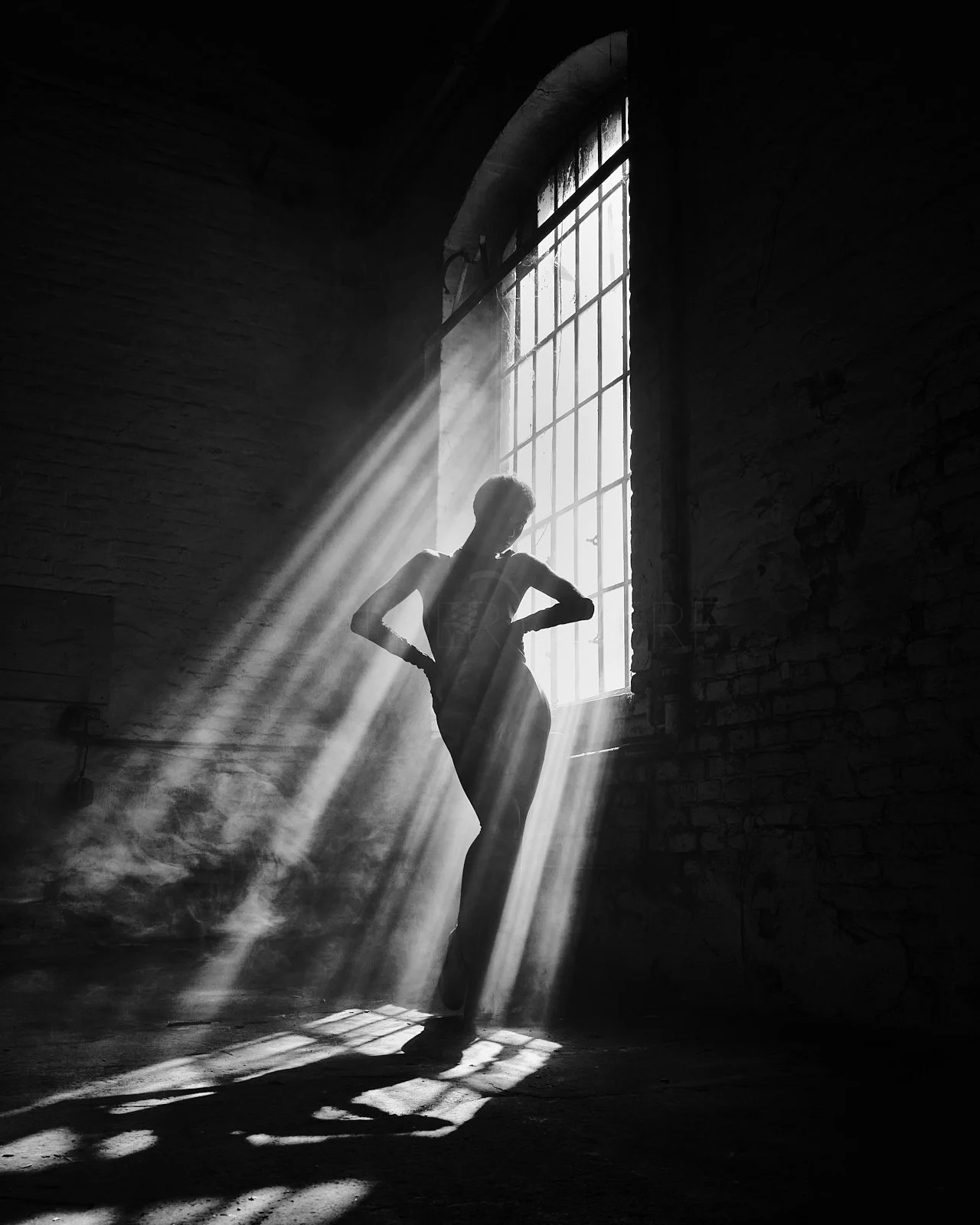A woman posing in front of a large window with sunlight streaming in, casting shadows in a dark, industrial space.