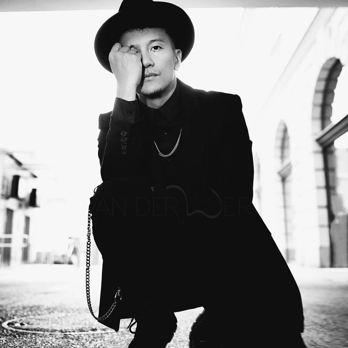 A black and white photo of a young man wearing a hat and dark clothing, squatting outdoors with a hand covering one eye, in an urban setting.