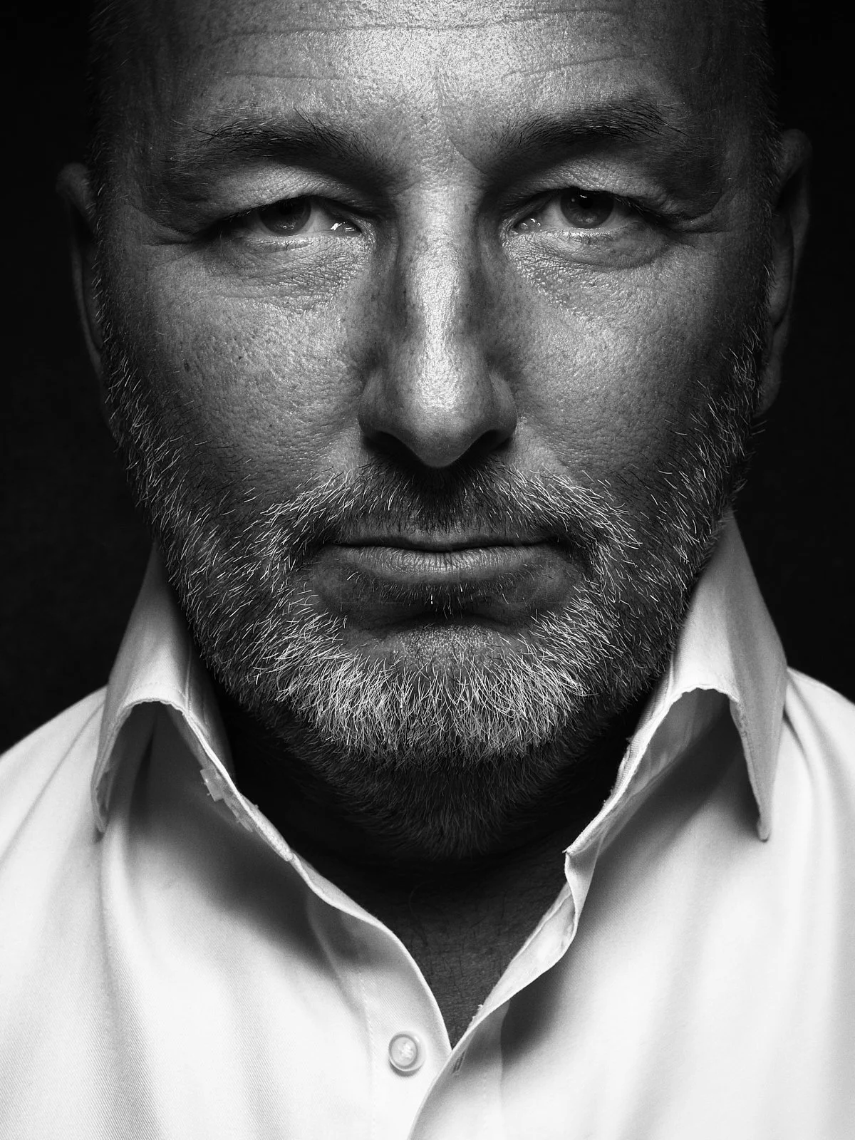 Black and white close-up portrait of a middle-aged man with a beard, wearing a collared shirt, looking directly at the camera with a serious expression.