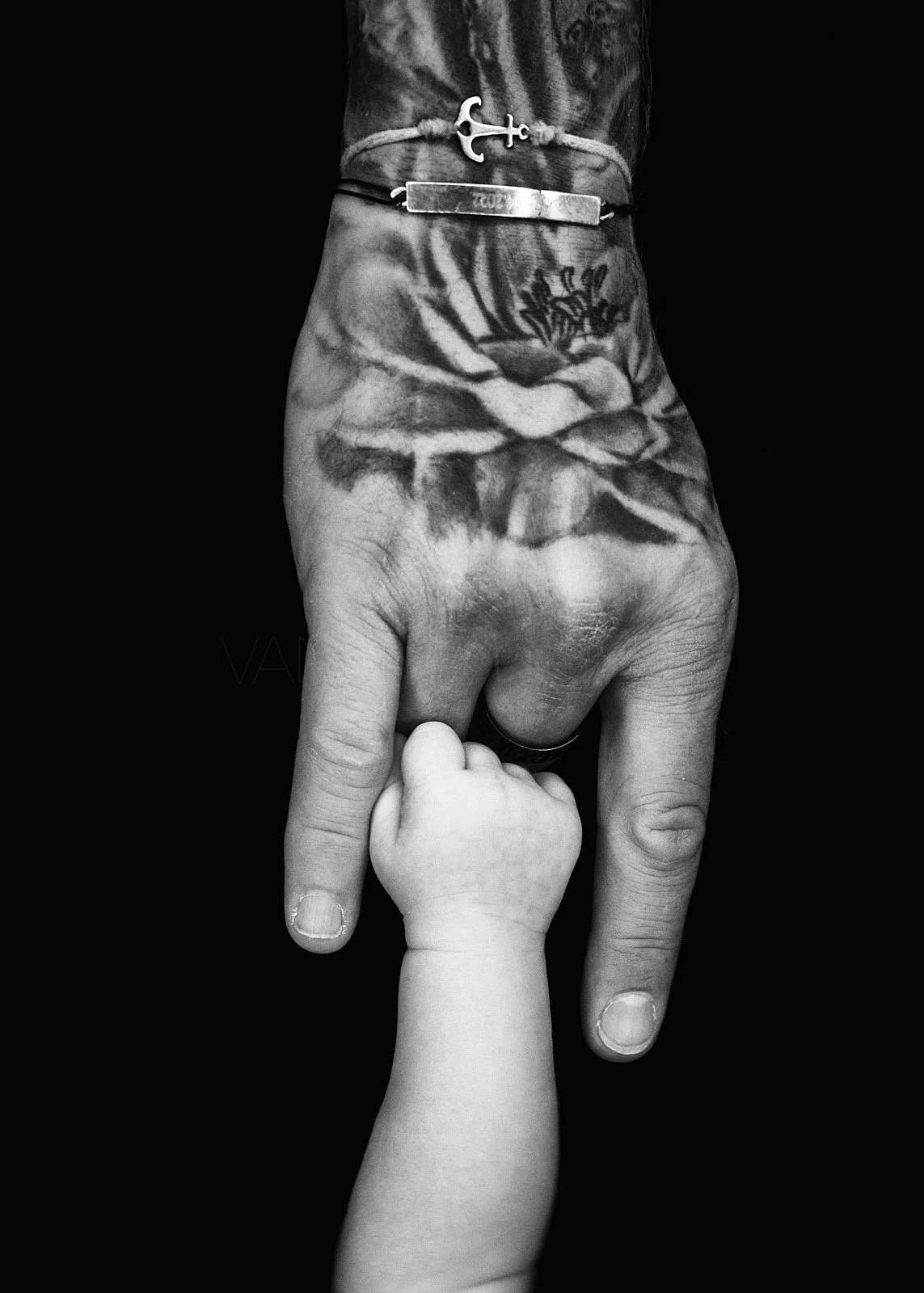 Black and white photo of a tattooed adult hand holding a small child's hand against a dark background.