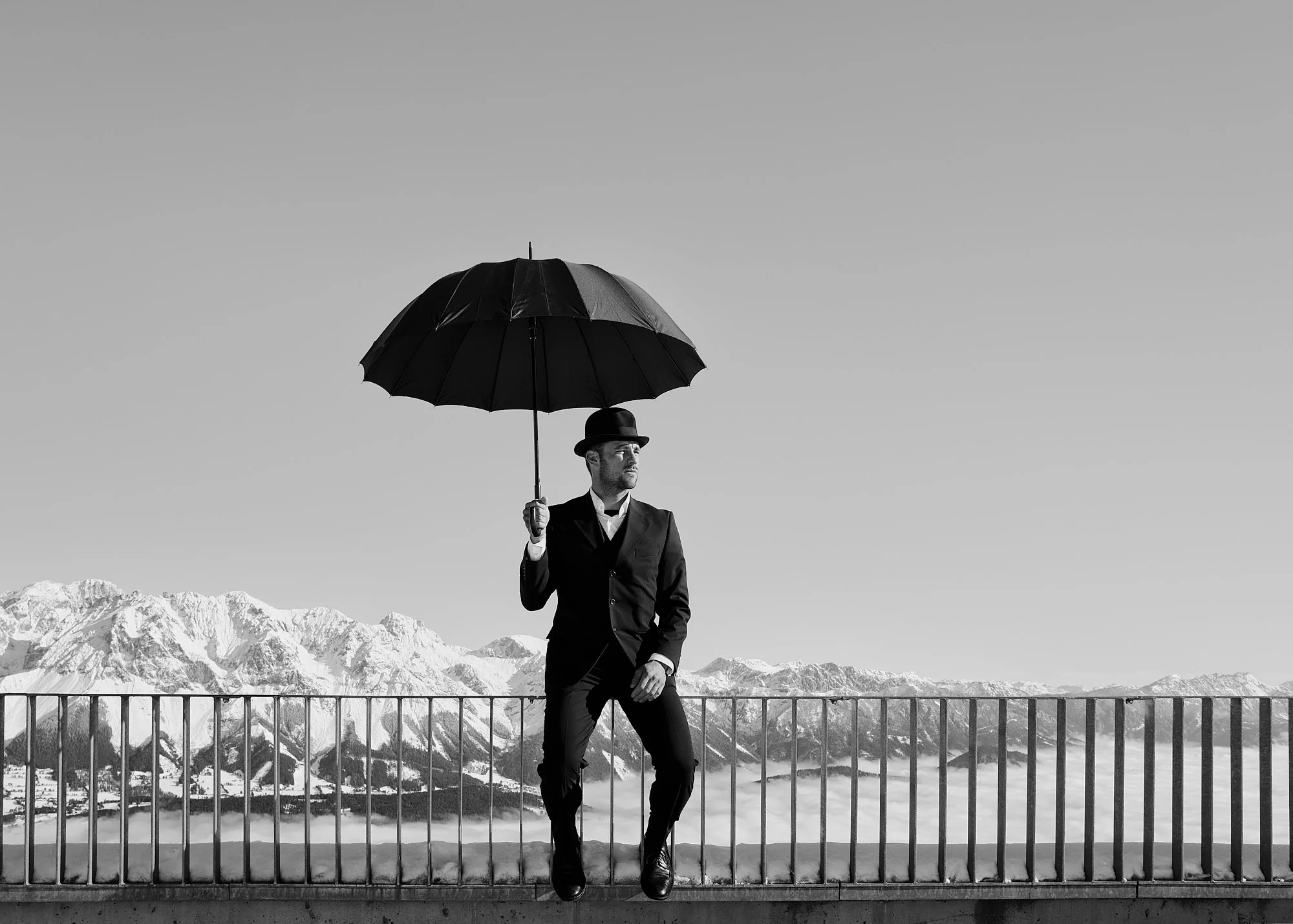 A man in a suit and bowler hat holding an umbrella, sitting on a railing against a mountain backdrop in black and white.