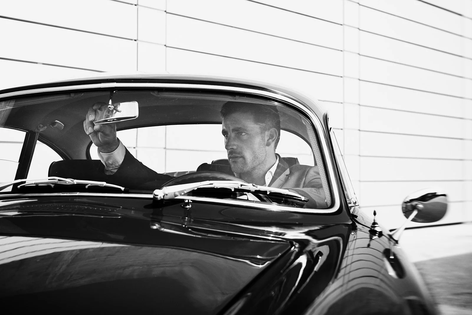 A man in a suit driving a classic black vintage car, adjusting the rearview mirror, with a plain white wall in the background.