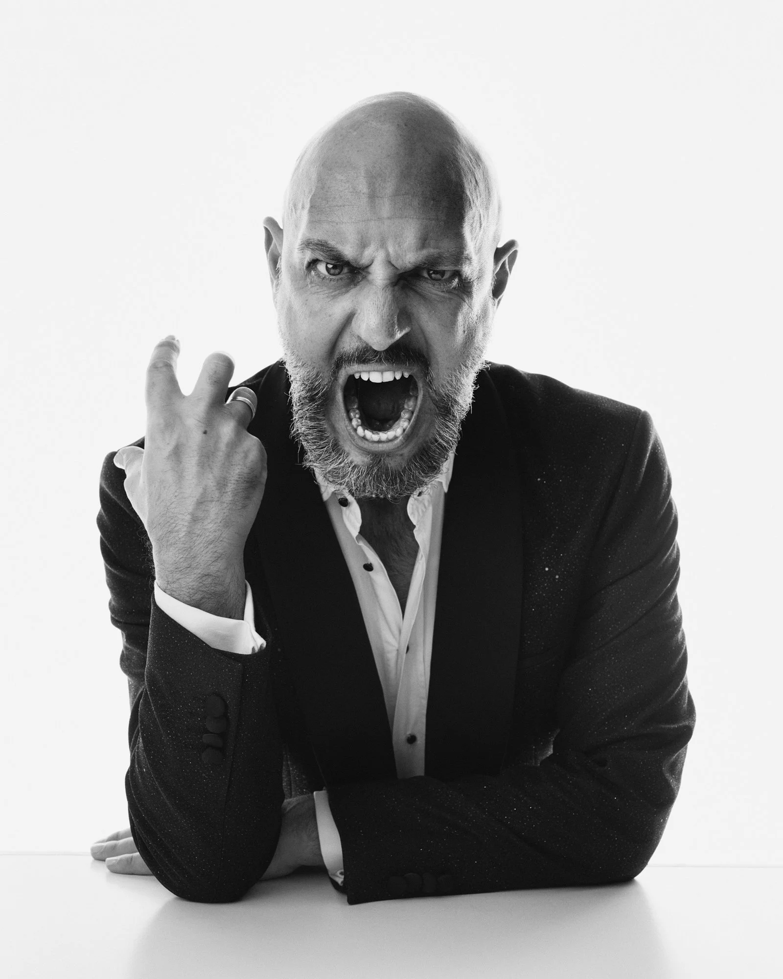 Black and white photo of a bald man with a beard, shouting with an angry expression, wearing a suit, with one hand clenched in a fist near his face.