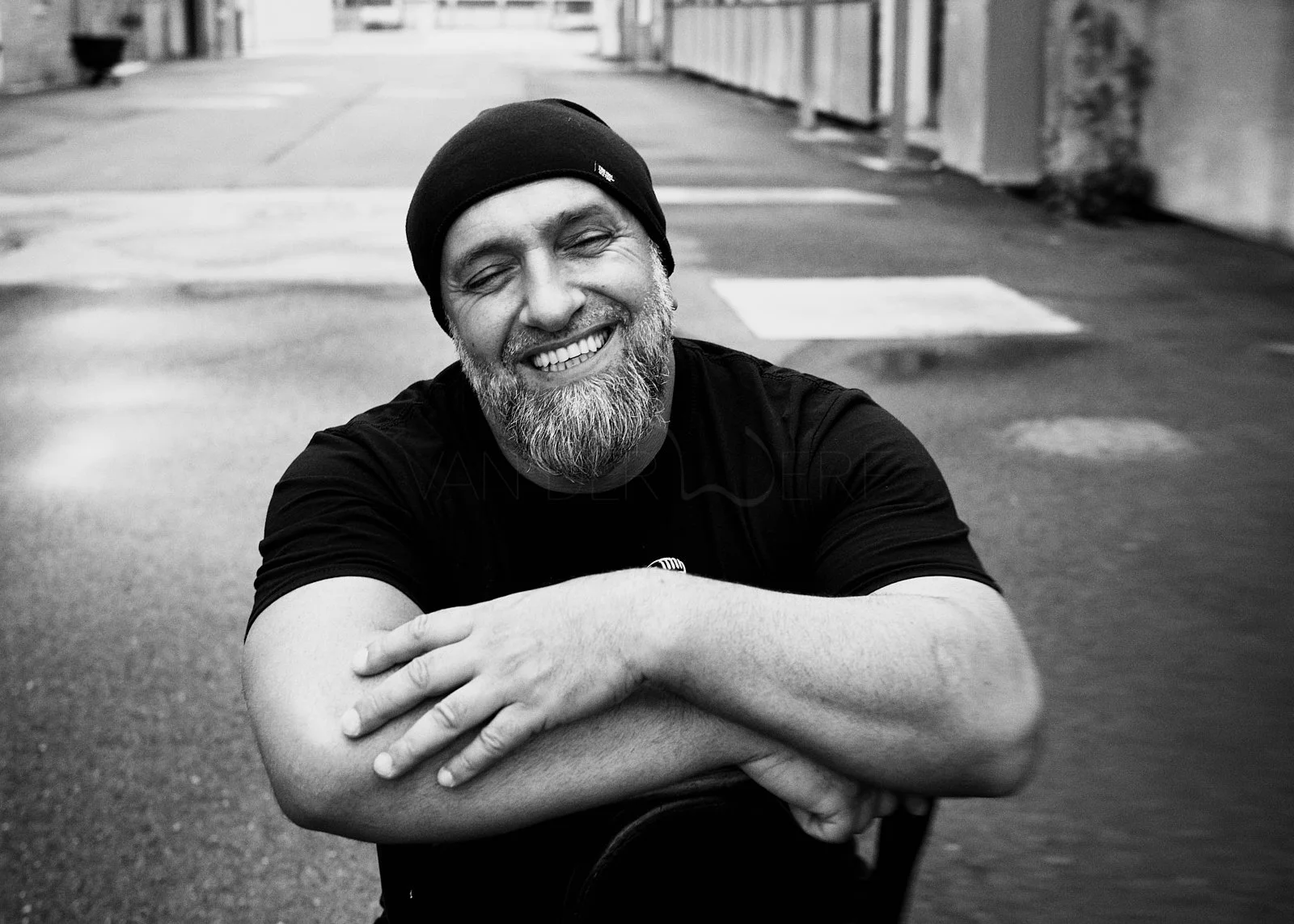 Smiling man with beard wearing a beanie and black t-shirt, sitting outdoors on an urban street with arms crossed, in black and white.