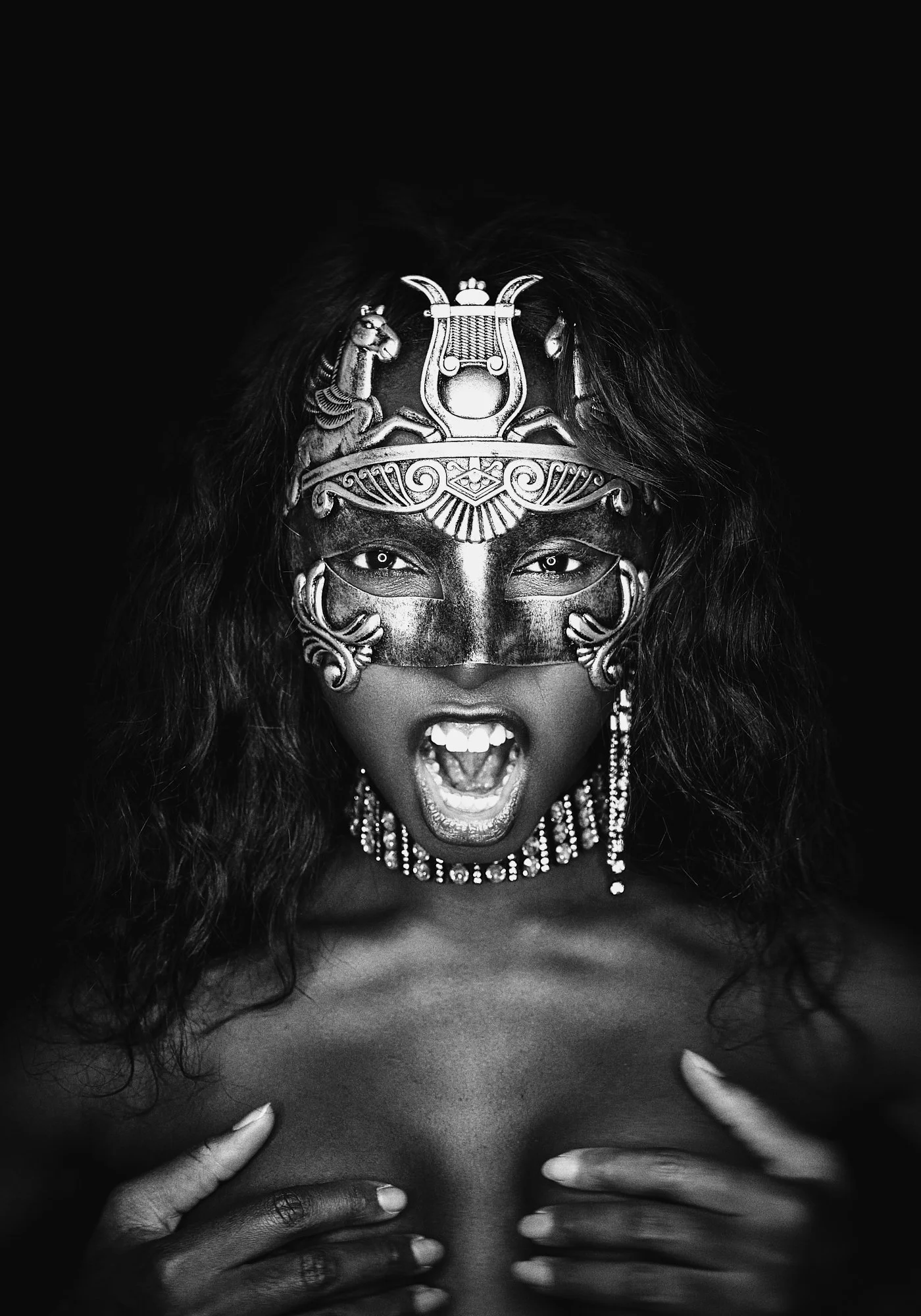 A woman wearing an ornate face mask with an Egyptian motif, jewelry, and her hands covering her chest, with her mouth open as if shouting, in black and white.