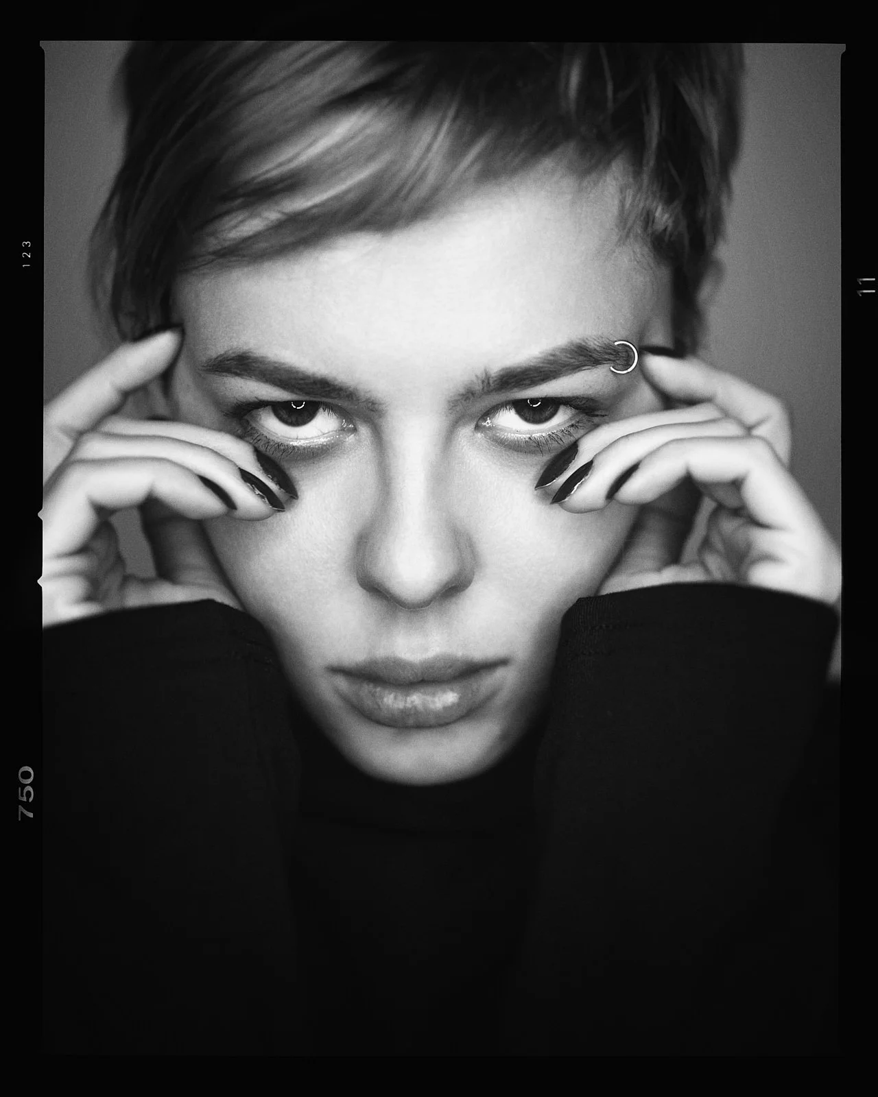 Black and white close-up portrait of a woman with short hair, intense gaze, and piercing eyes, holding her face with her hands. She has a nose ring and dark nail polish.