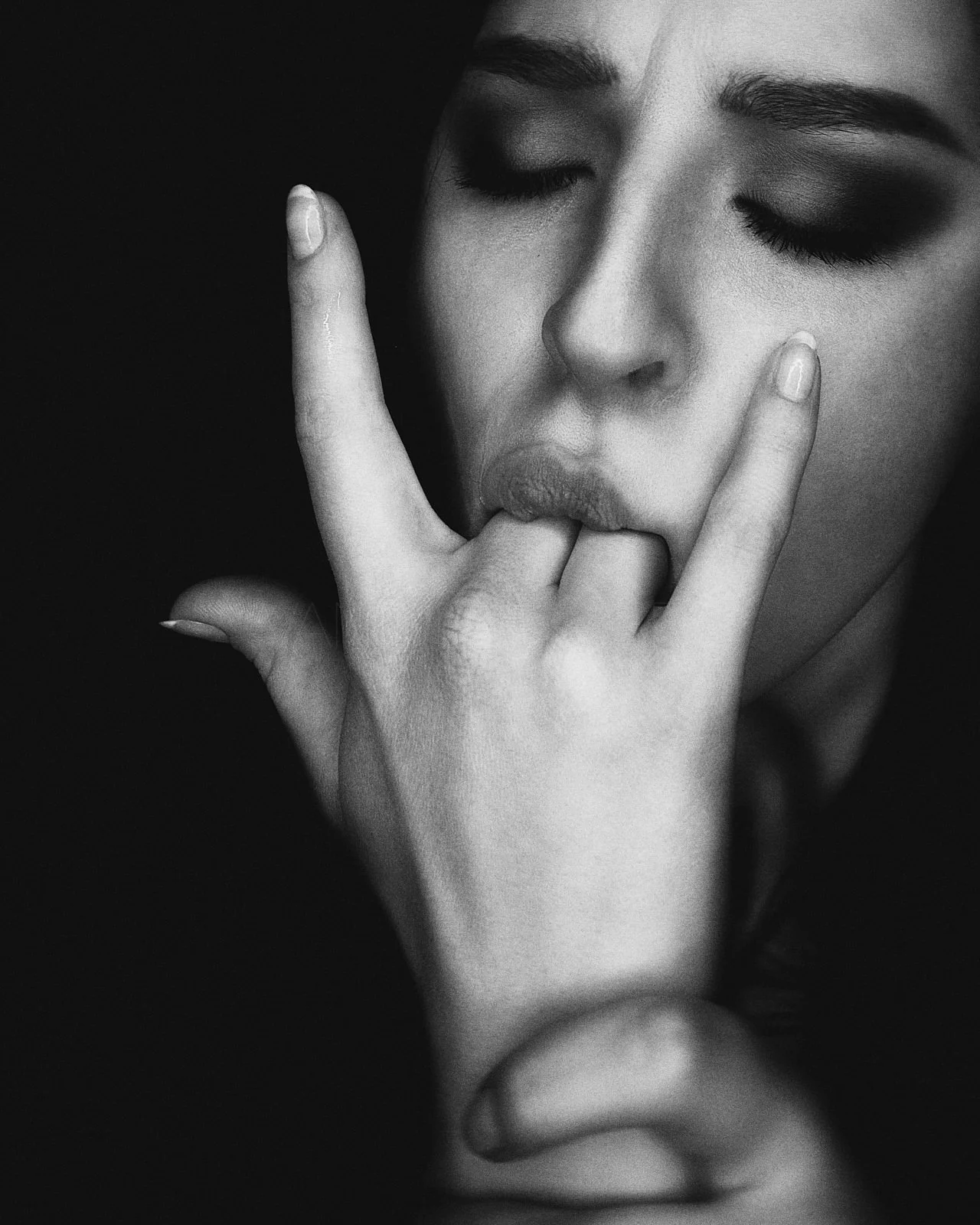 A black and white close-up photo of a woman with her eyes closed, lips slightly pursed, and fingers in a 'rock on' gesture near her mouth.