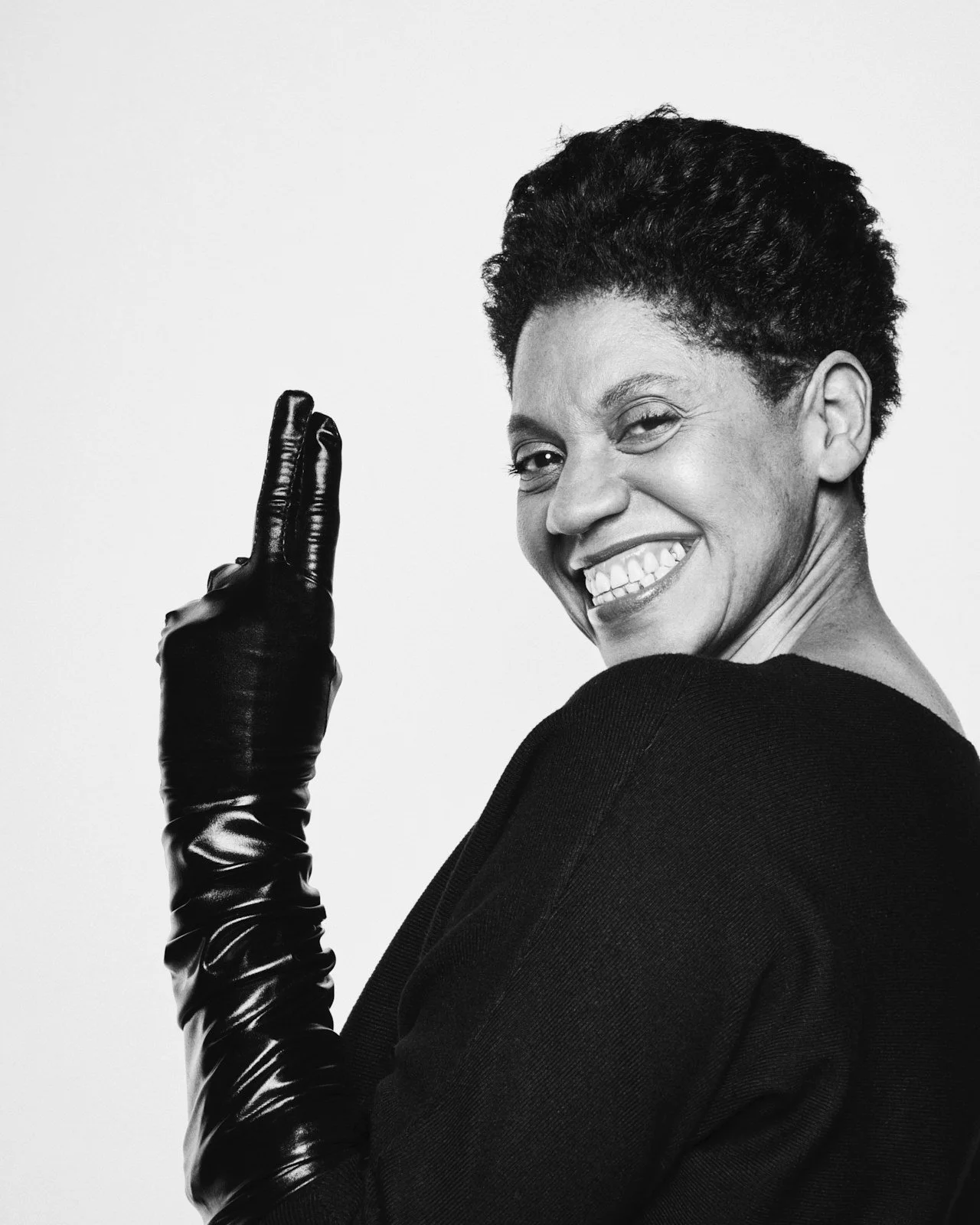 A smiling woman with short curly hair wearing a black top and a black leather glove on her right hand, making a peace sign against a plain background.