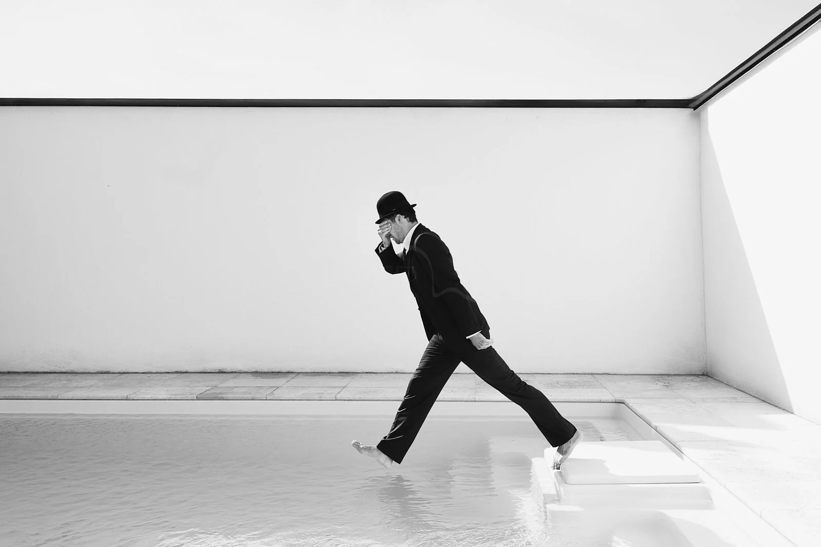 A man dressed in a suit and bowler hat wades into a shallow pool in a minimalist, white room with geometric shadows, walking with a contemplative posture.
