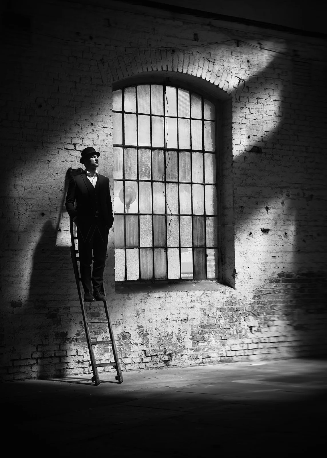 A man in a suit and hat standing on a ladder against a brick wall near a large window with metal bars, with dramatic lighting creating shadows in an industrial-style space.