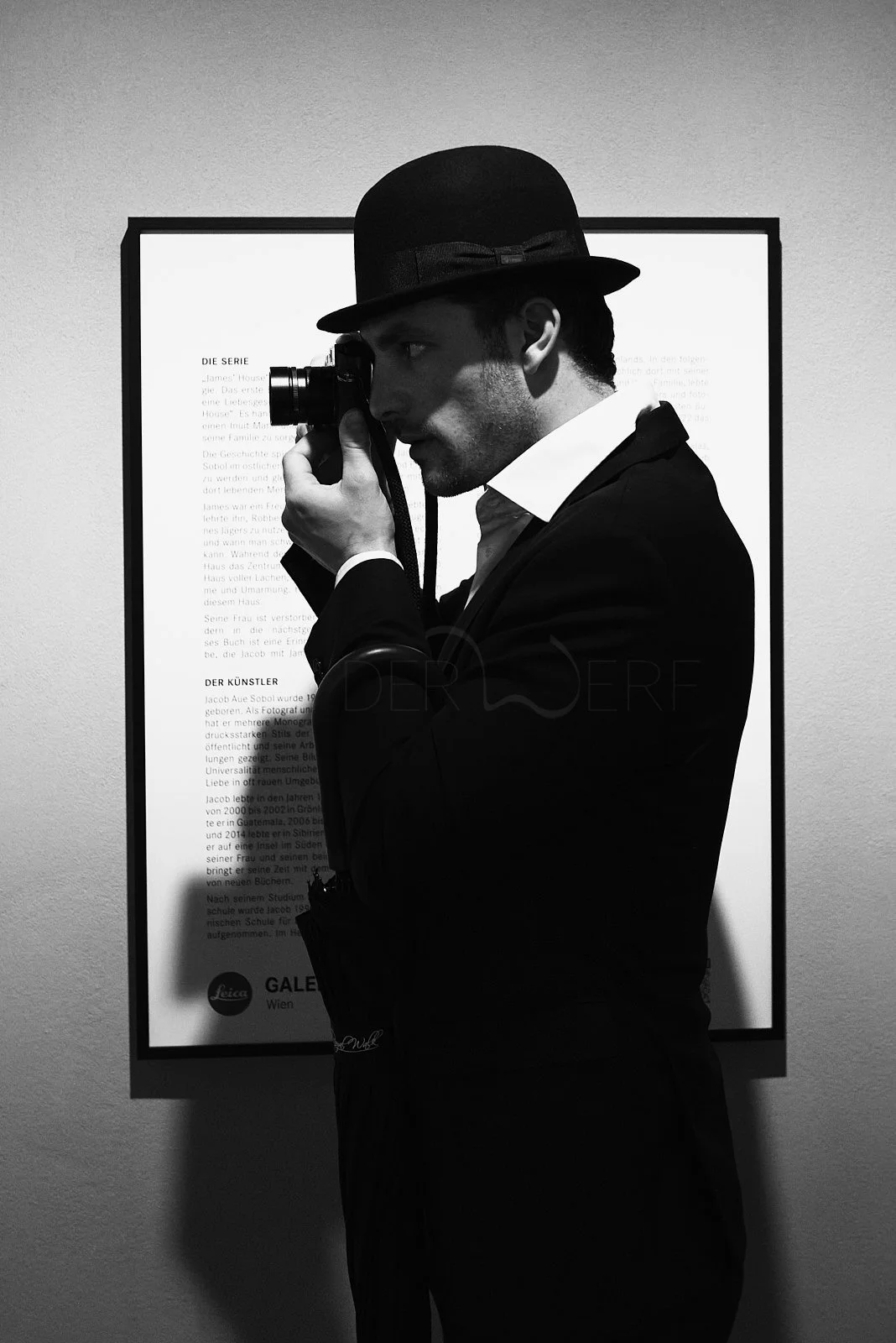 Man wearing a hat and suit, taking a photograph with a vintage camera in an art gallery, with a framed display behind him.