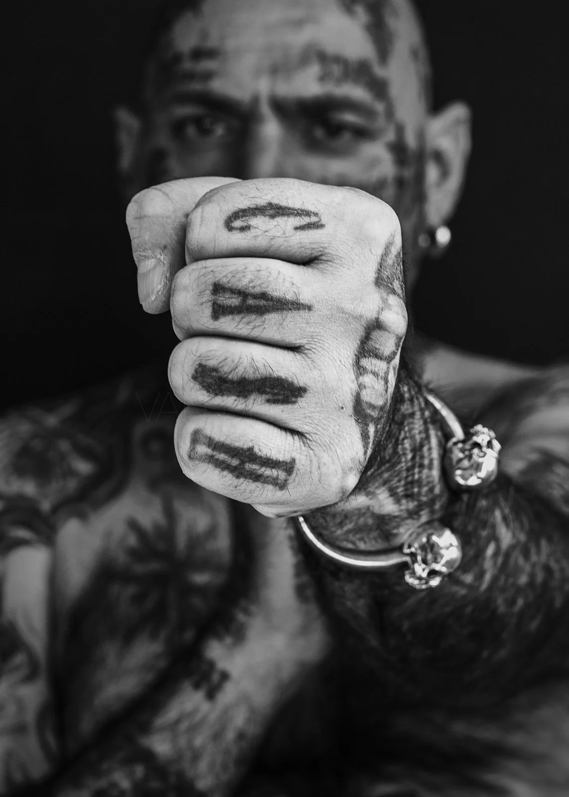 Close-up of a tattooed man's clenched fist with graffiti-style writing, pointing towards the camera, with tattoos on his face and arm, wearing a chain necklace with skull charms, in black and white.