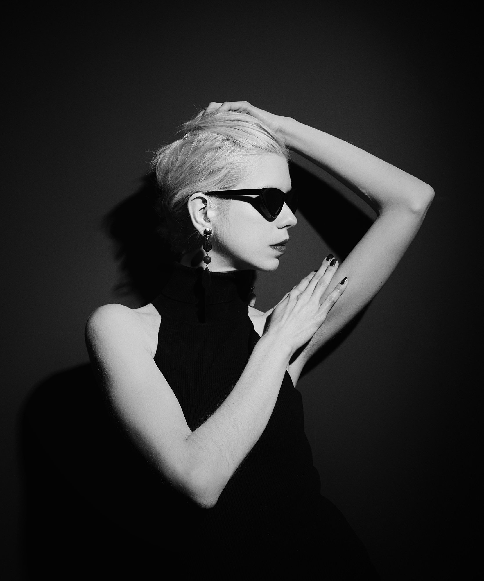 Black and white portrait of a woman wearing sunglasses, with one arm raised, and jewelry, against a dark background.